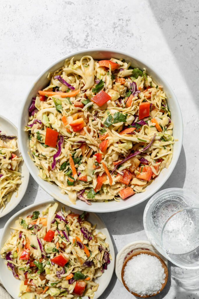 a white bowl of southwestern style coleslaw next to two white plates of slaw salad.