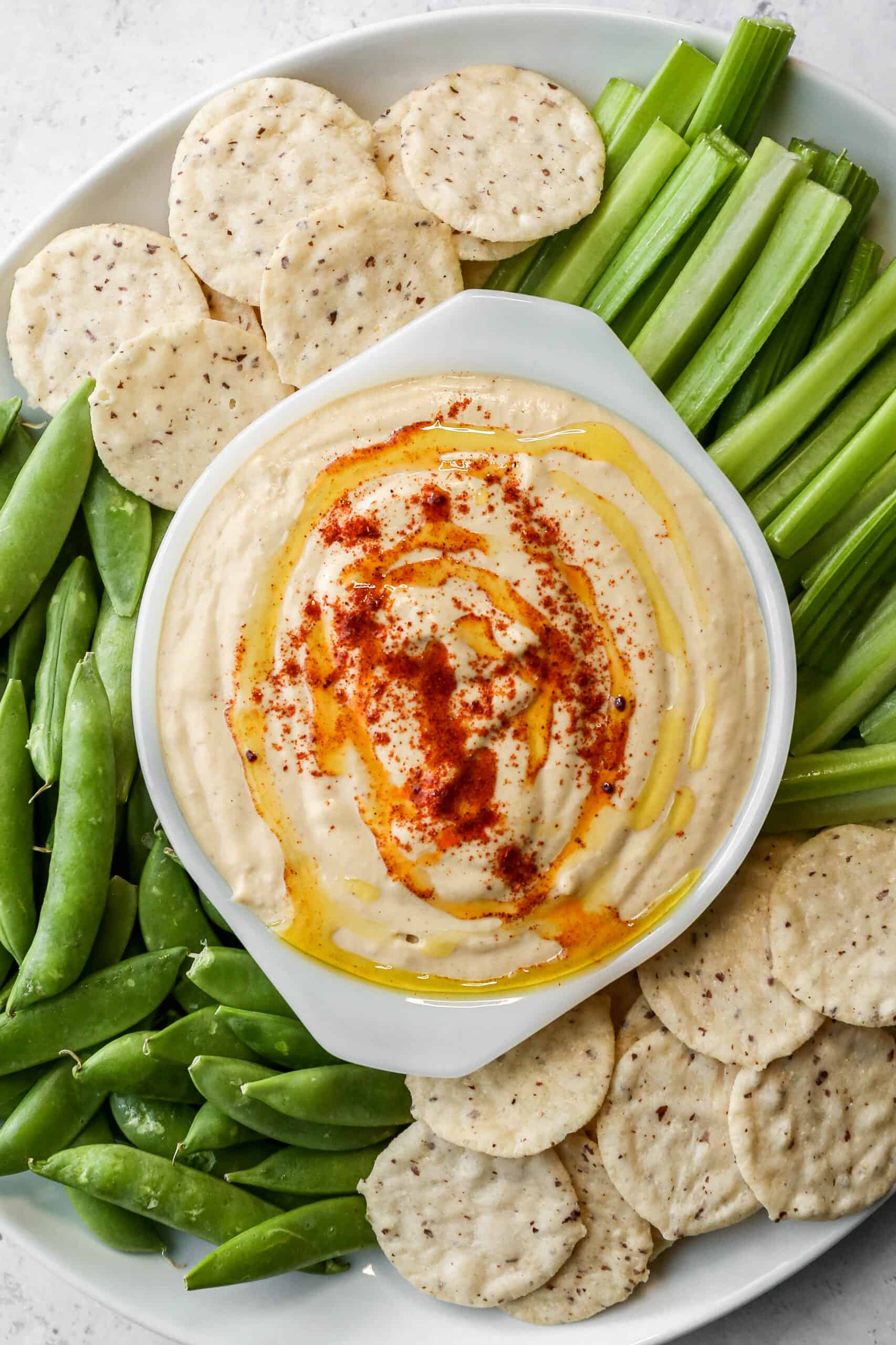 a small white bowl of gluten free easy homemade garlic hummus surrounded by celery sticks, sugar snap peas and rice crackers.