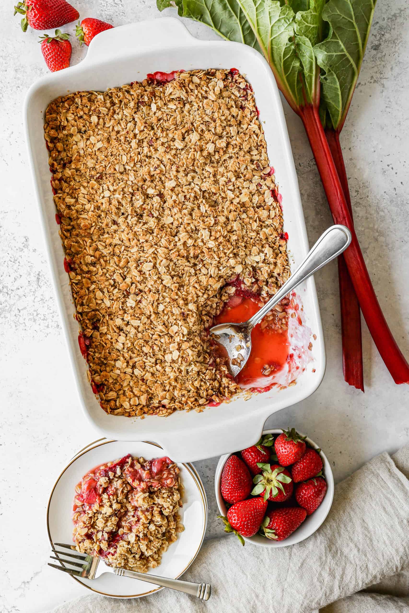 a white 9x13 baking dish of gluten free strawberry rhubarb crumble next to stalks of fresh rhubarb and a small bowl of fresh strawberries.