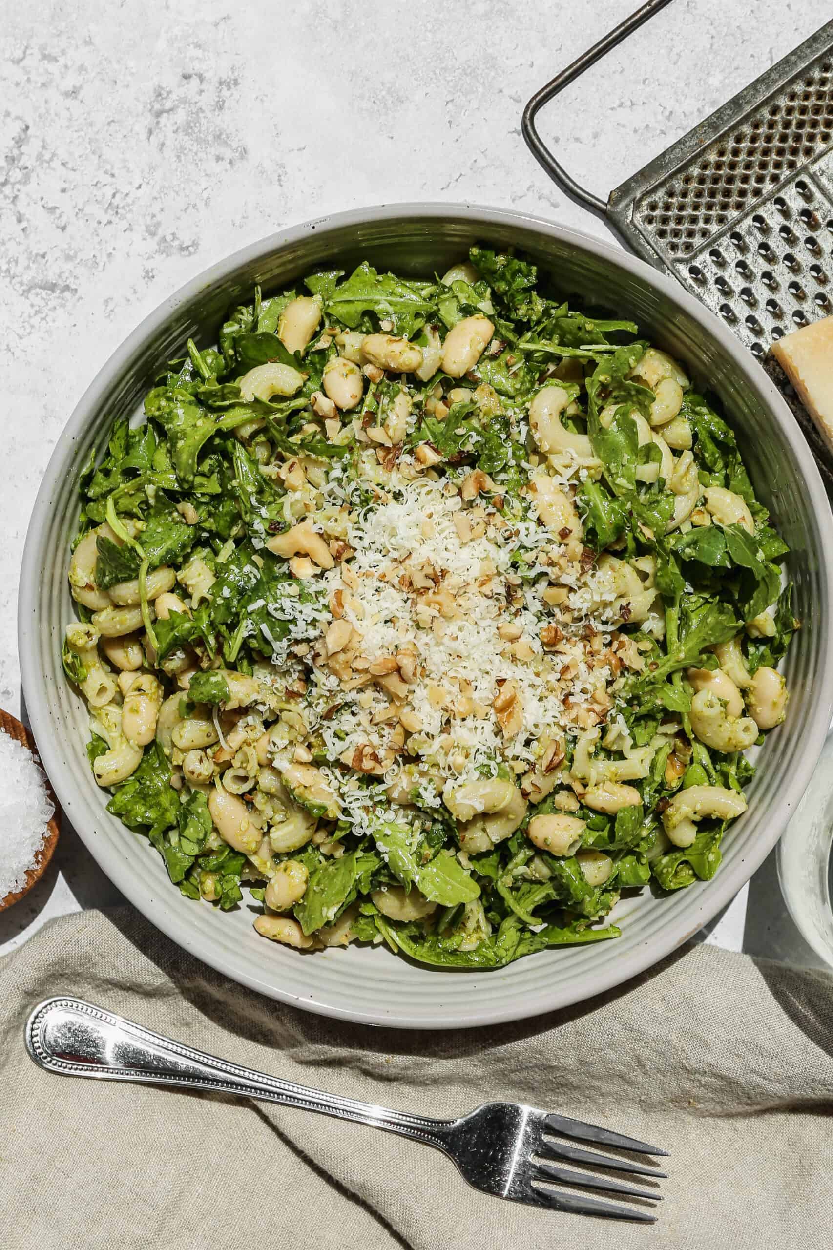 a white bowl of gluten free pesto pasta salad next to a grater with a small block of parmesan cheese on it.