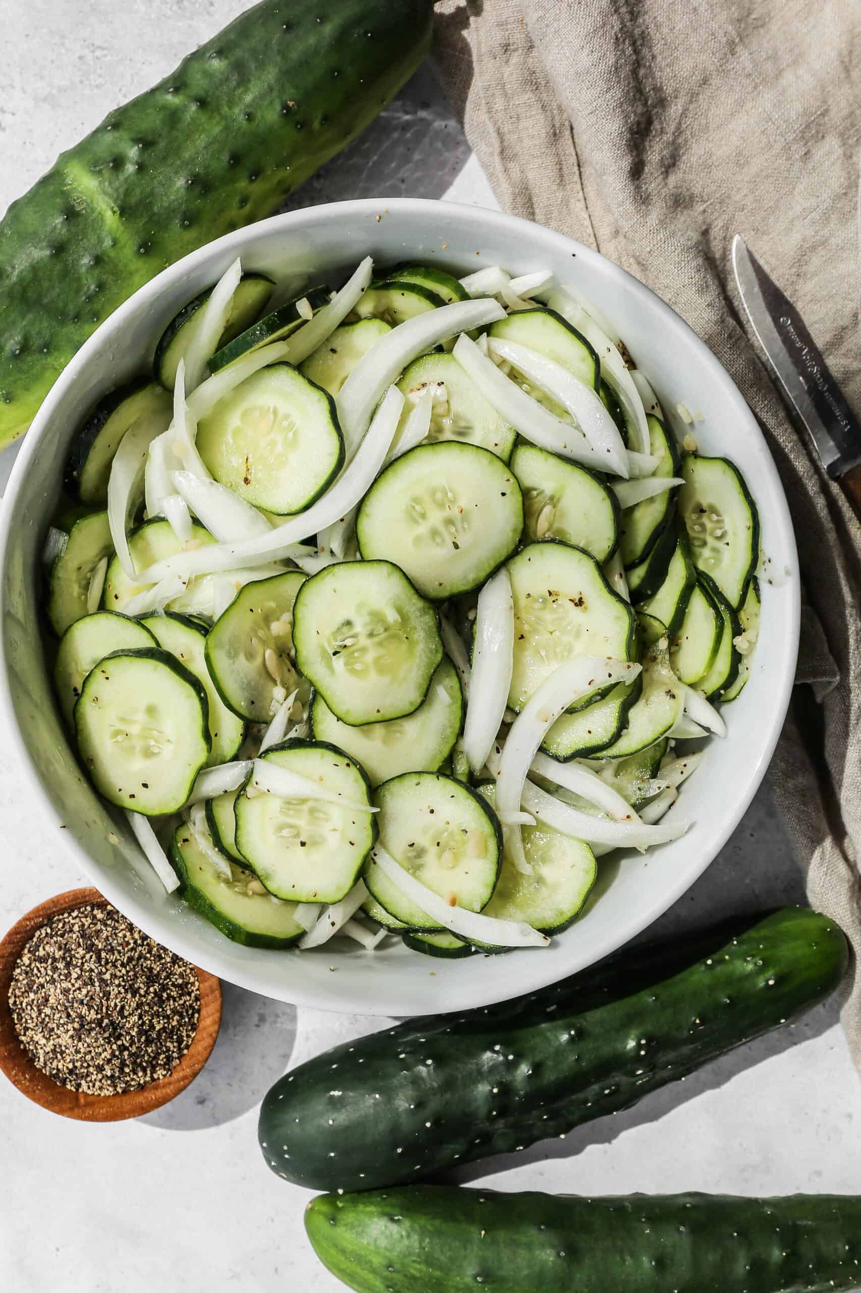 a white bowl of cucumber salad with vinegar dressing next to fresh cucumbers and a small wooden bowl of coarse ground black pepper.