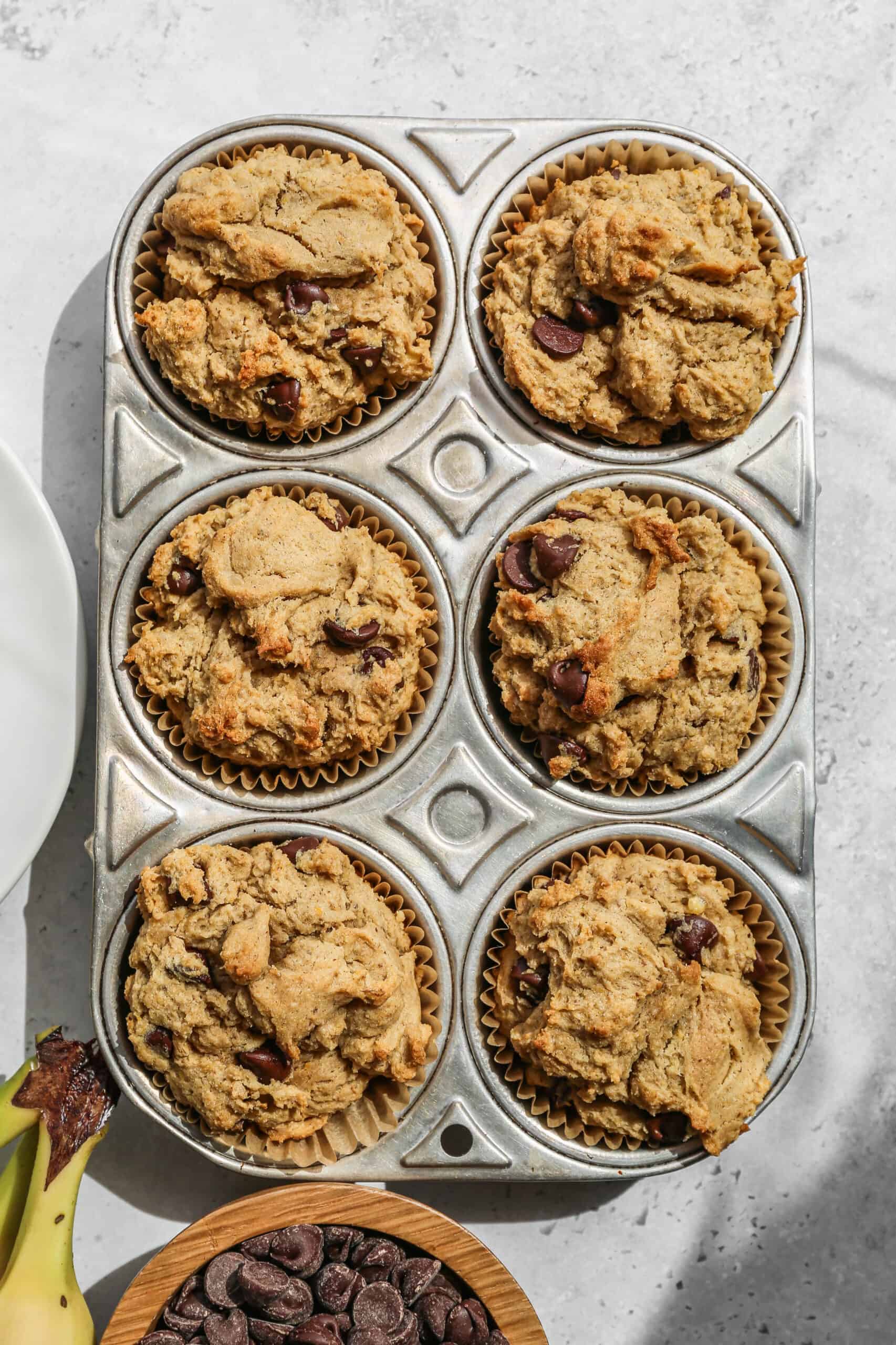 a metal muffin tin of gluten free peanut butter banana muffins next to a small wooden bowl of chocolate chips.