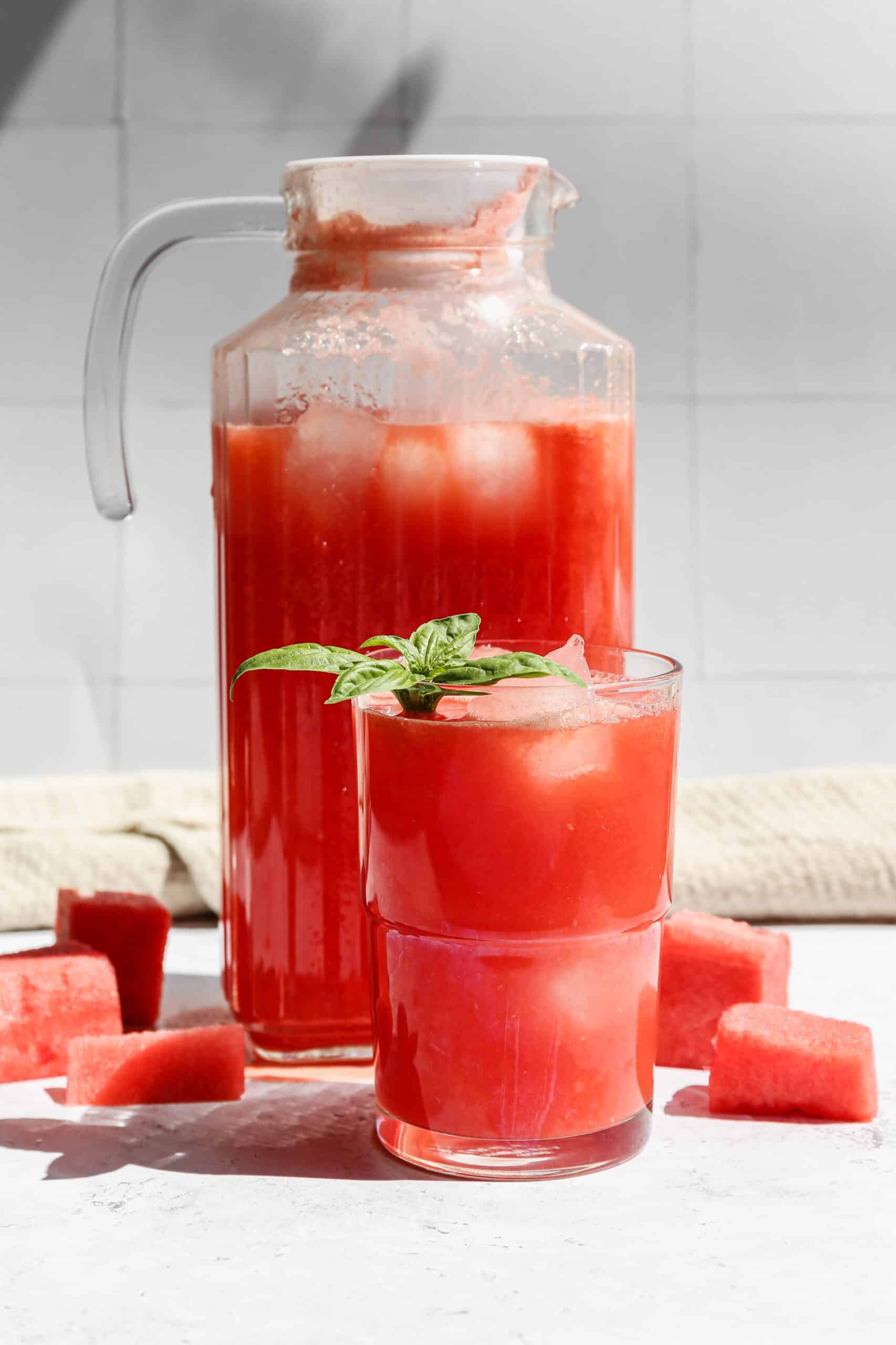 a glass pitcher and a glass tumbler of watermelon lemonade next to chunks of fresh watermelon.