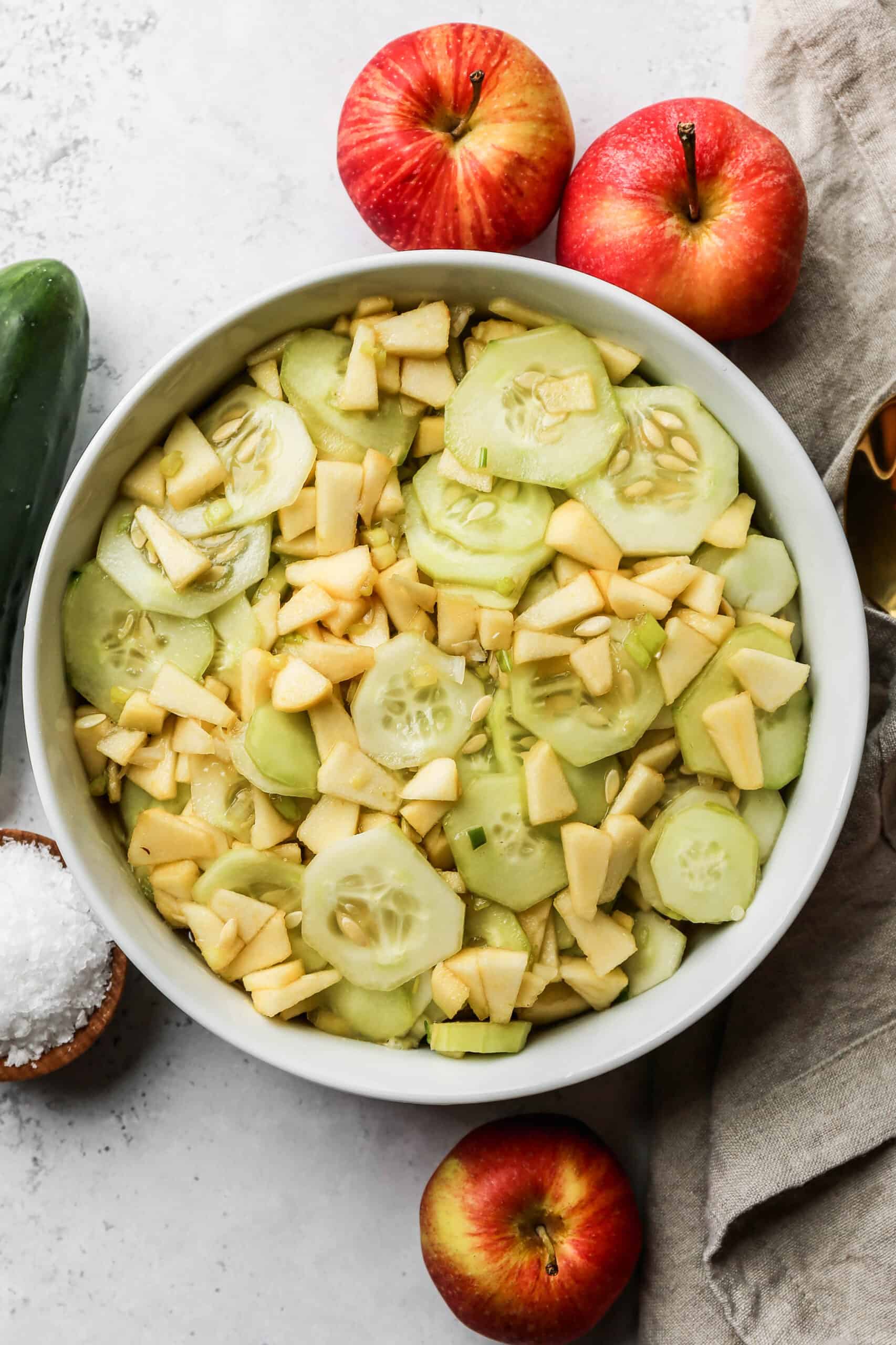a white serving bowl of apple cucumber salad next to fresh apples and cucumbers.