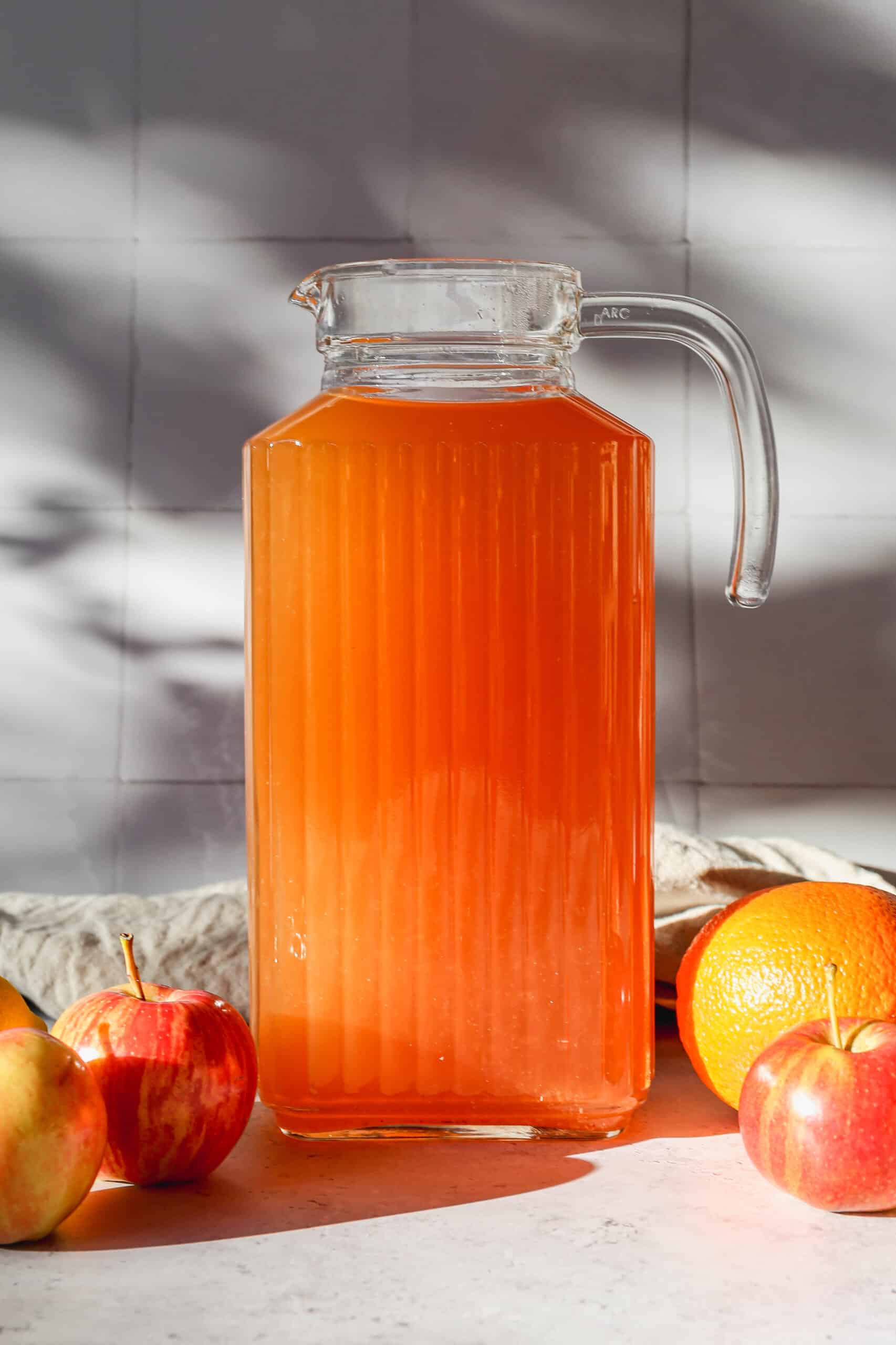 a glass pitcher of easy instant pot apple cider surrounded by fresh apples, oranges and lemon.