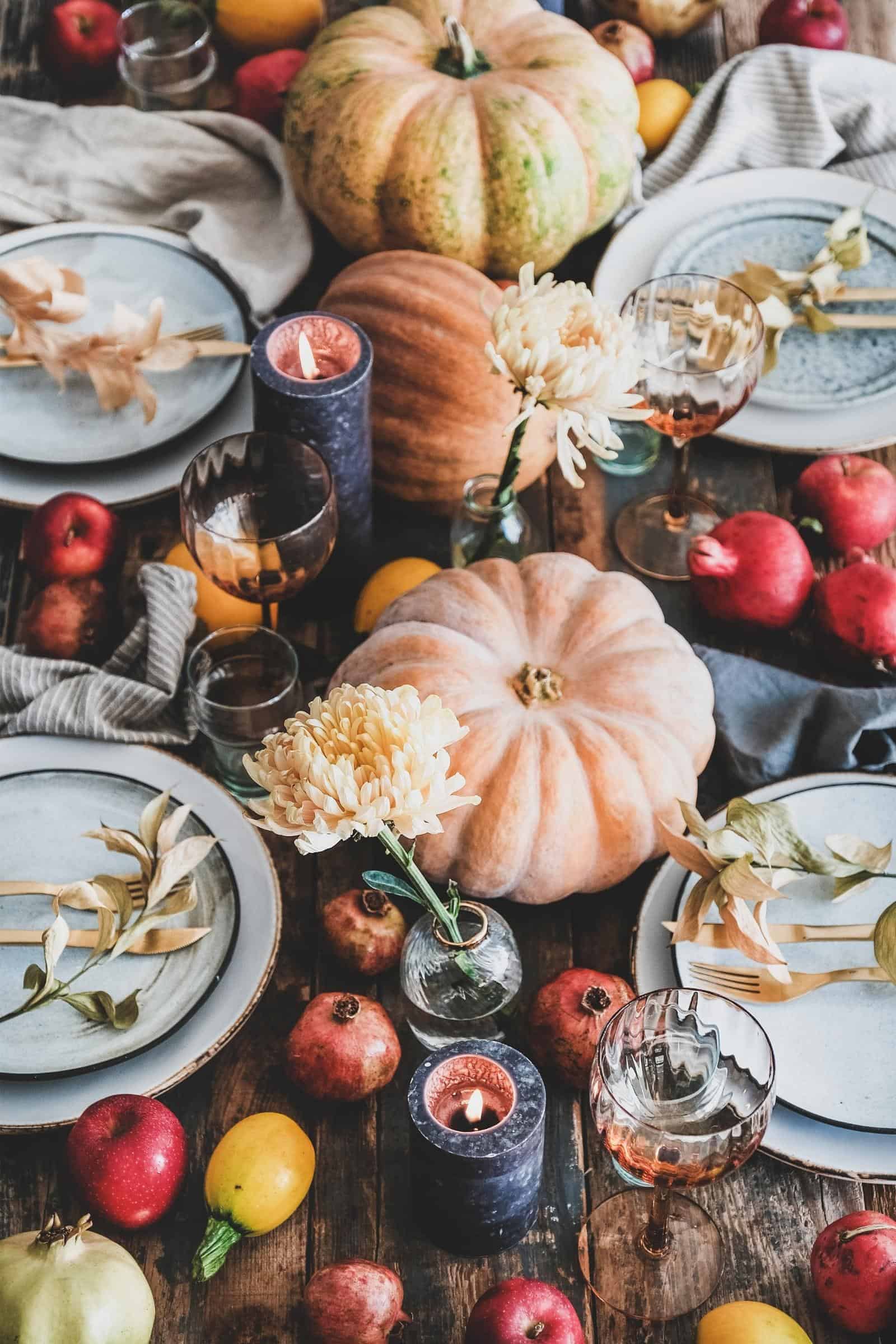 a Thanksgiving table set with plates, wine glasses, pumpkins, pomegranates, flowers and linen napkins.