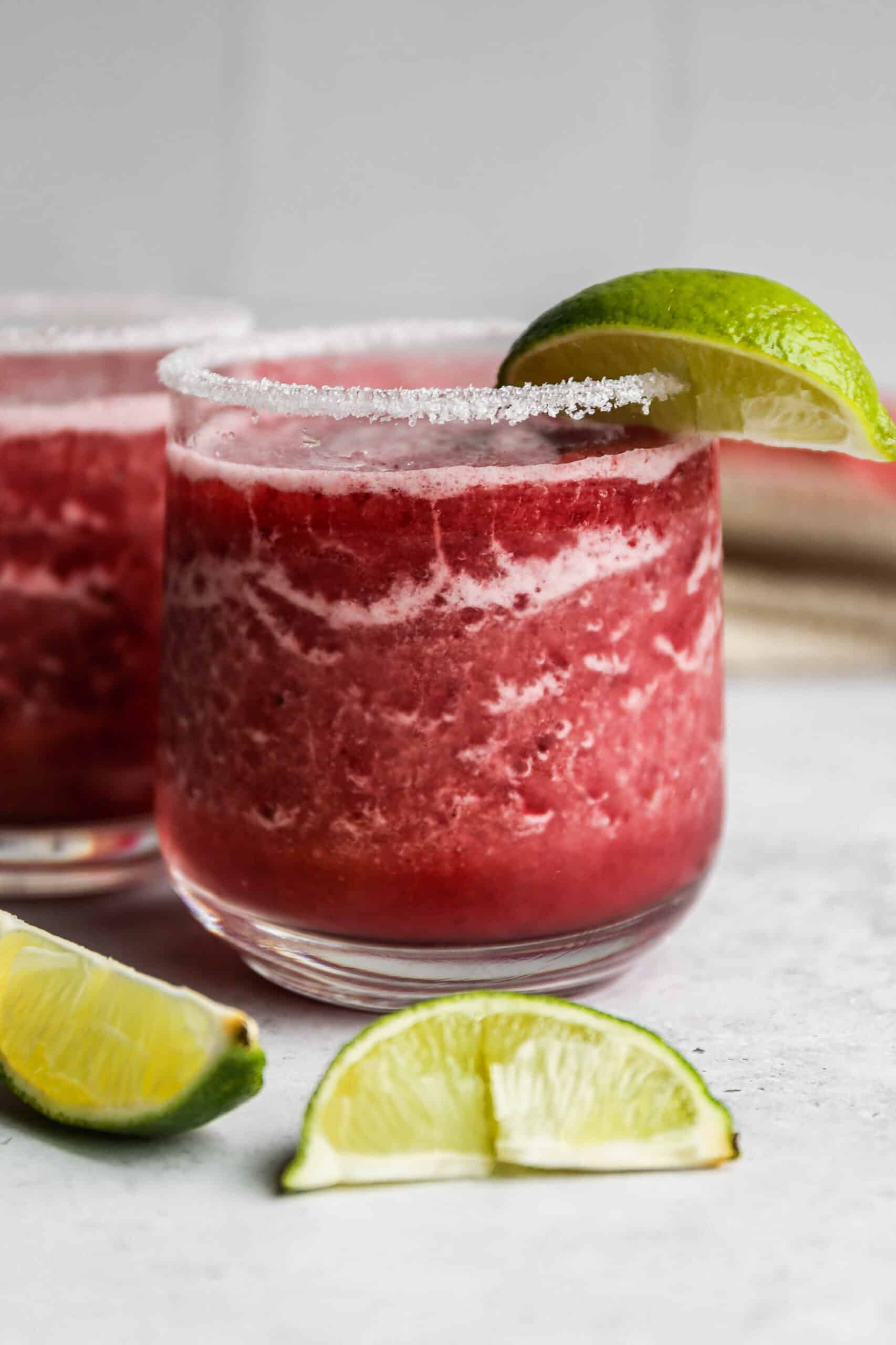 two glasses of frozen cranberry margarita mocktails with salt on the rim and a lime wedge.