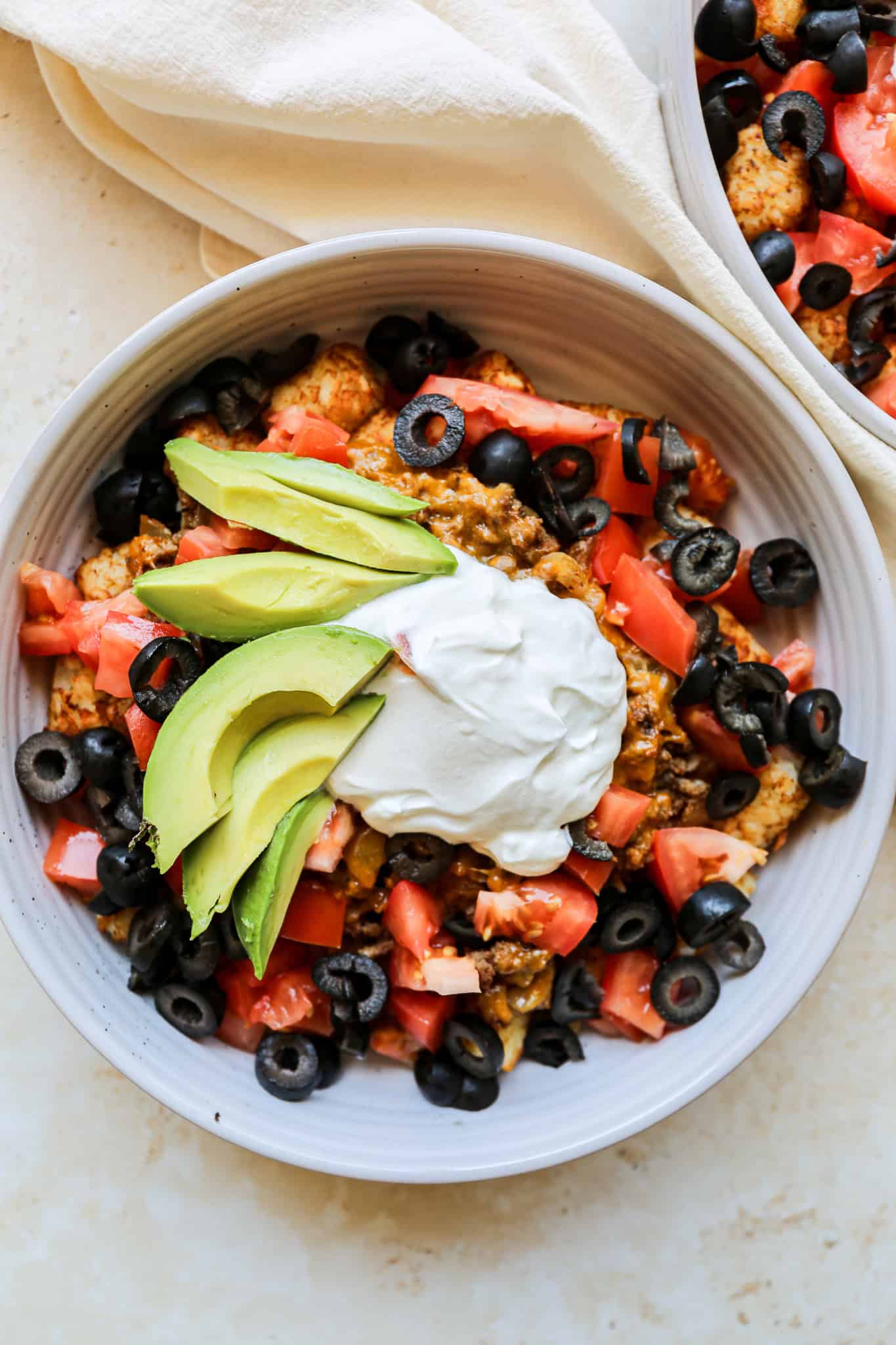 a white bowl with gluten free tater tot taco topped with sour cream, chopped tomatoes, diced black olives and sliced avocado