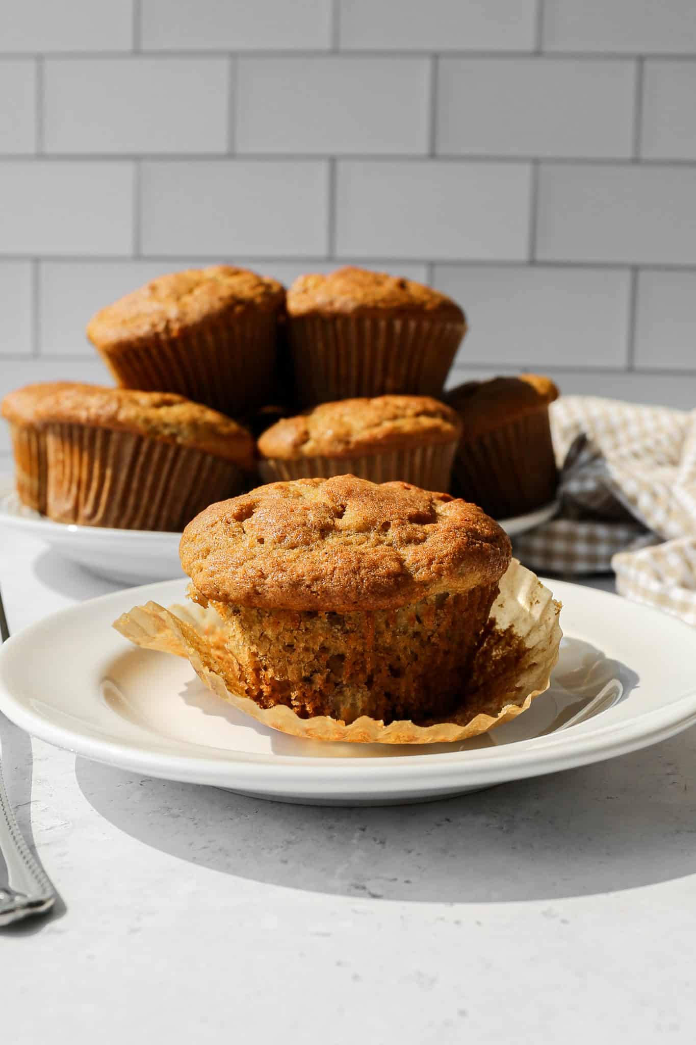 a gluten free banana bread muffin on a white plate with its liner wrapper pulled down with a plate of muffins sitting behind it