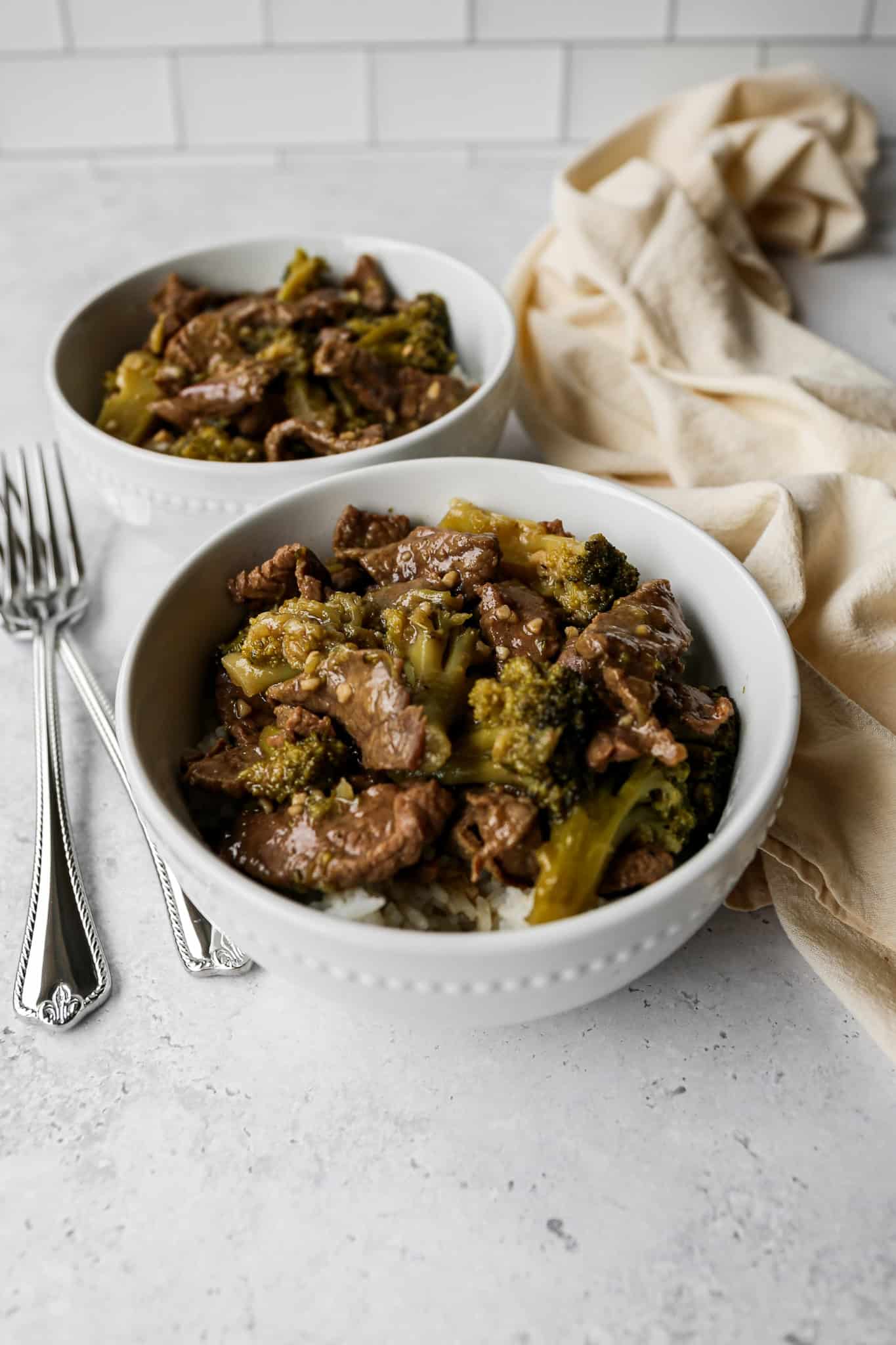 two white bowls of gluten free slow cooker beef and broccoli next to silver forks and a linen kitchen towel