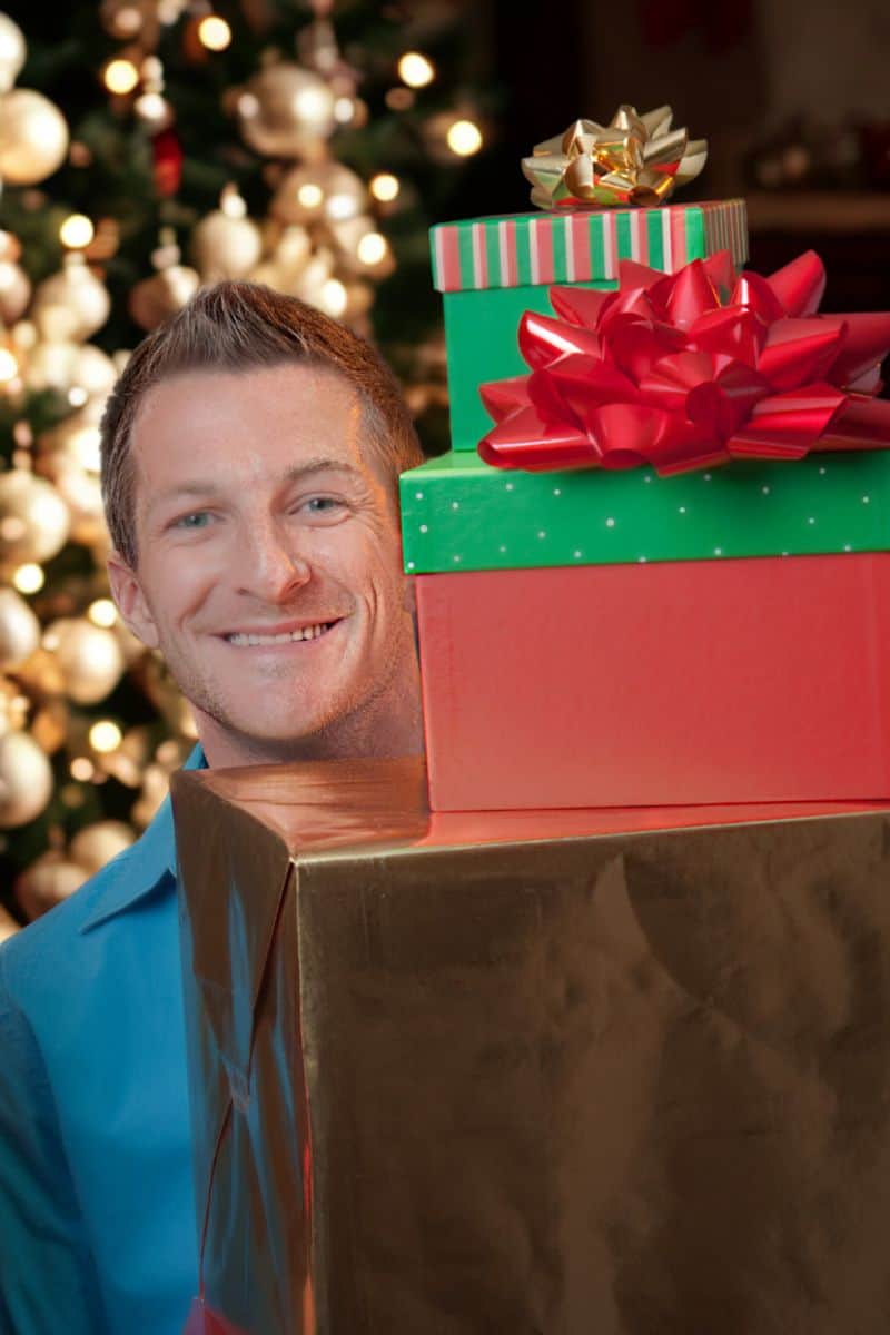 a man holding three large gifts with bows on top in front of a Christmas tree