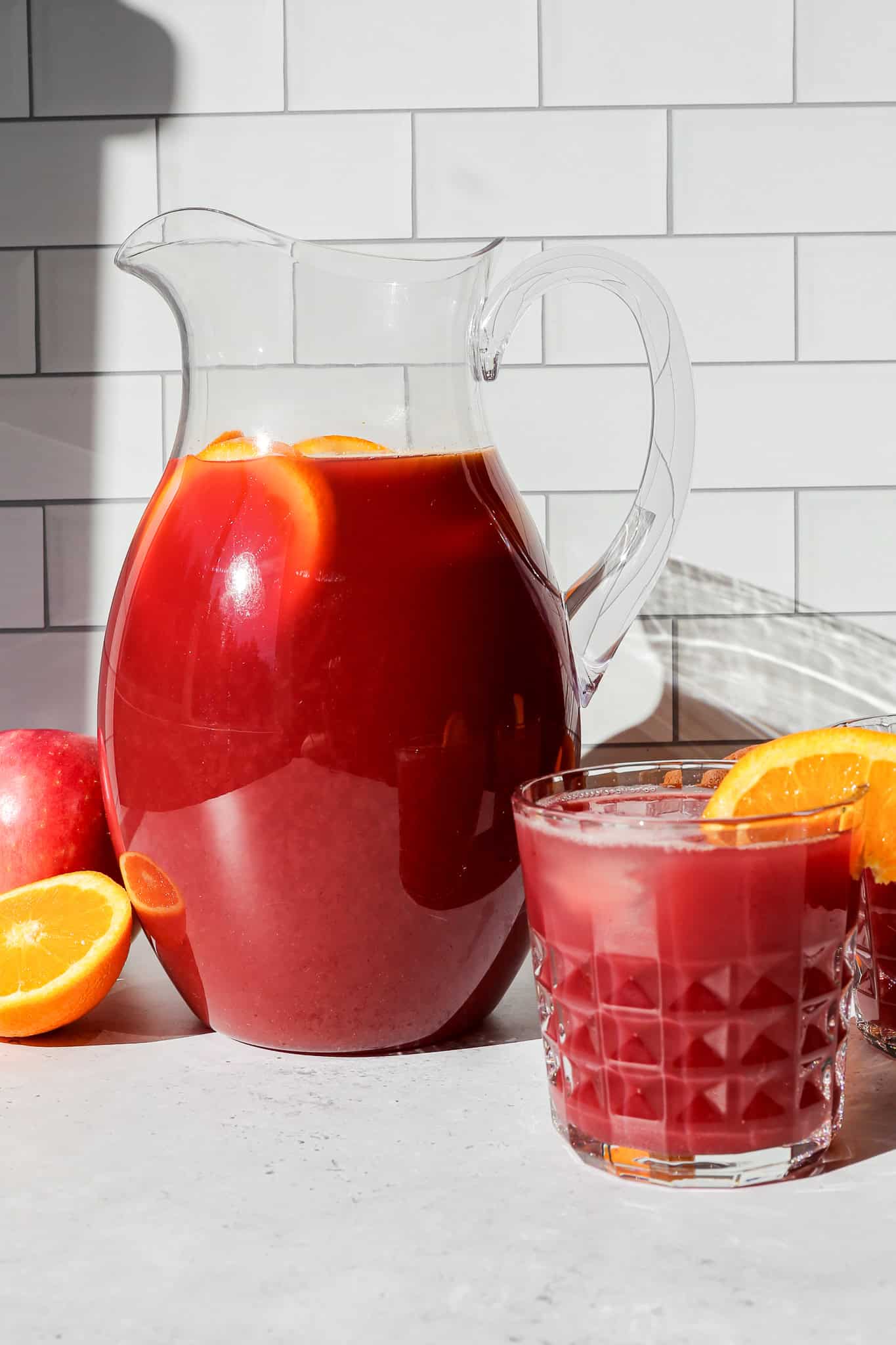 a clear pitcher of autumn harvest punch sitting next a drinking glass of the punch with an orange wedge on the rim