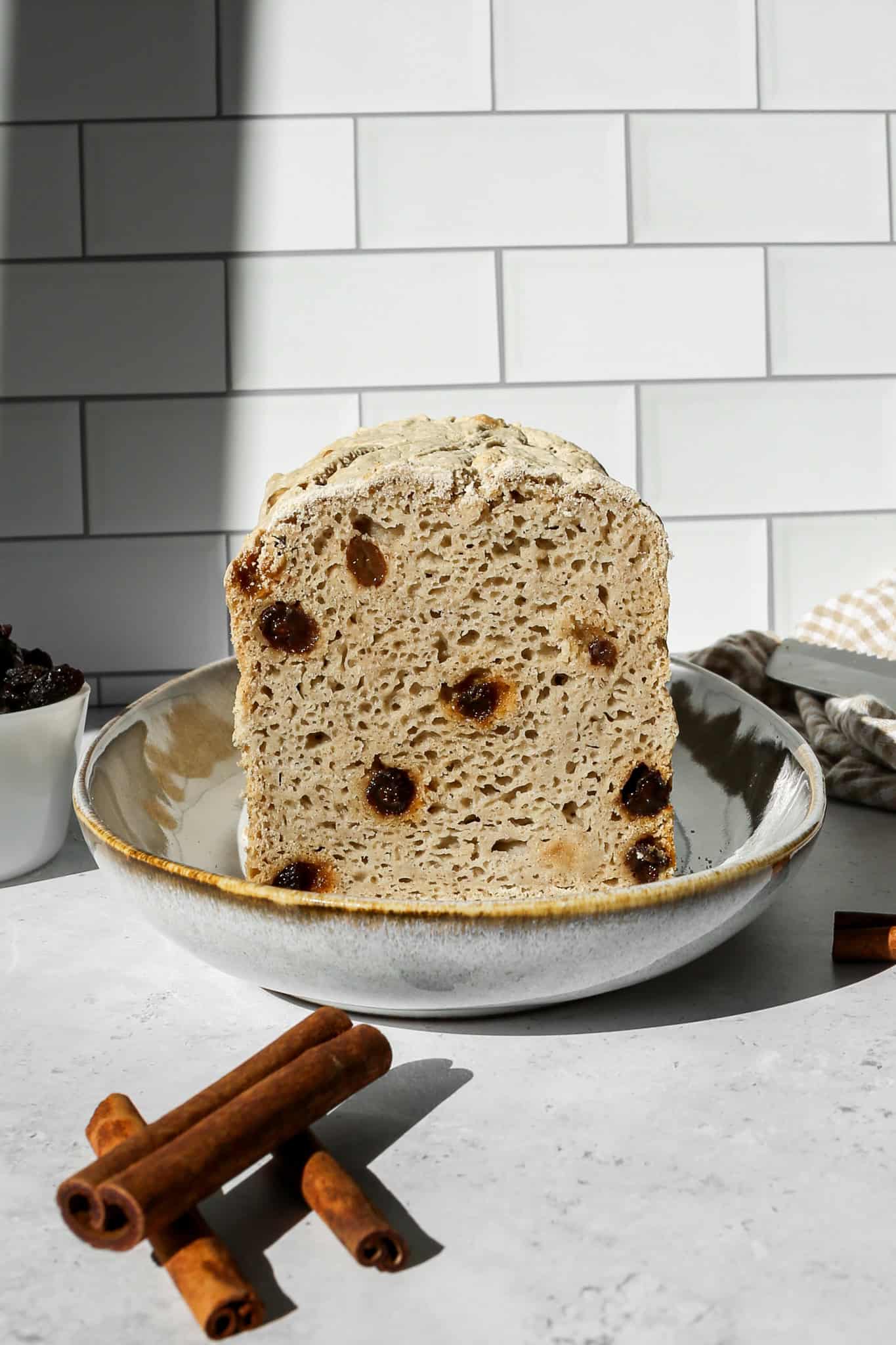 a loaf of gluten free cinnamon raisin sourdough bread on an oval platter next to a small white bowl of raisins, cinnamon sticks and a tan and white gingham kitchen towel