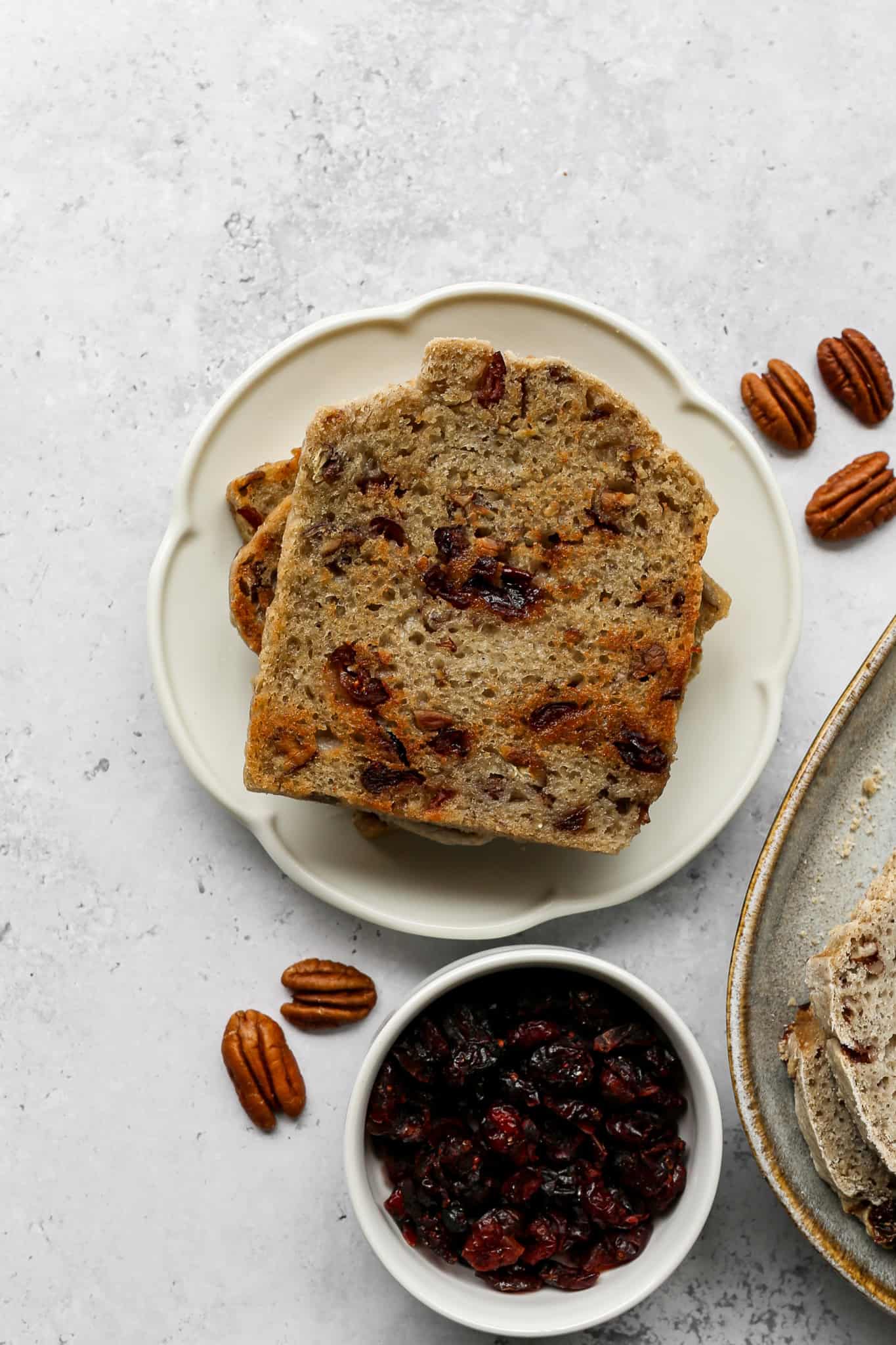 Toasted slices of gluten free cranberry pecan sourdough bread on a plate next to whole pecans and a small white bowl of dried cranberries
