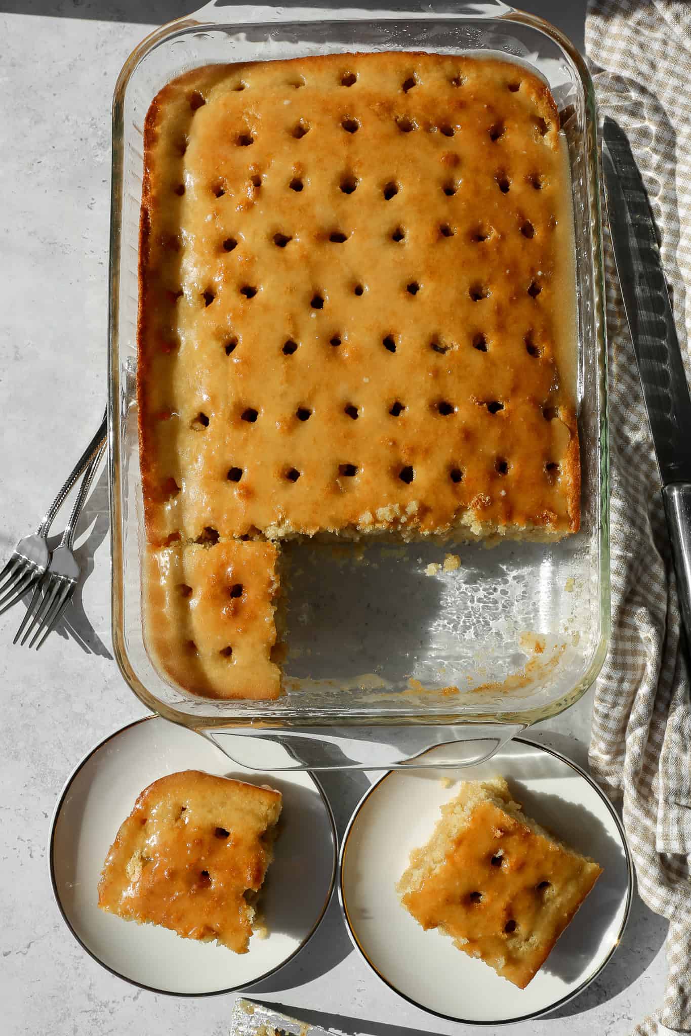 a glass 9x13 baking dish of gluten free vanilla buttermilk poke cake with two slice cut and on white plates sitting next to it