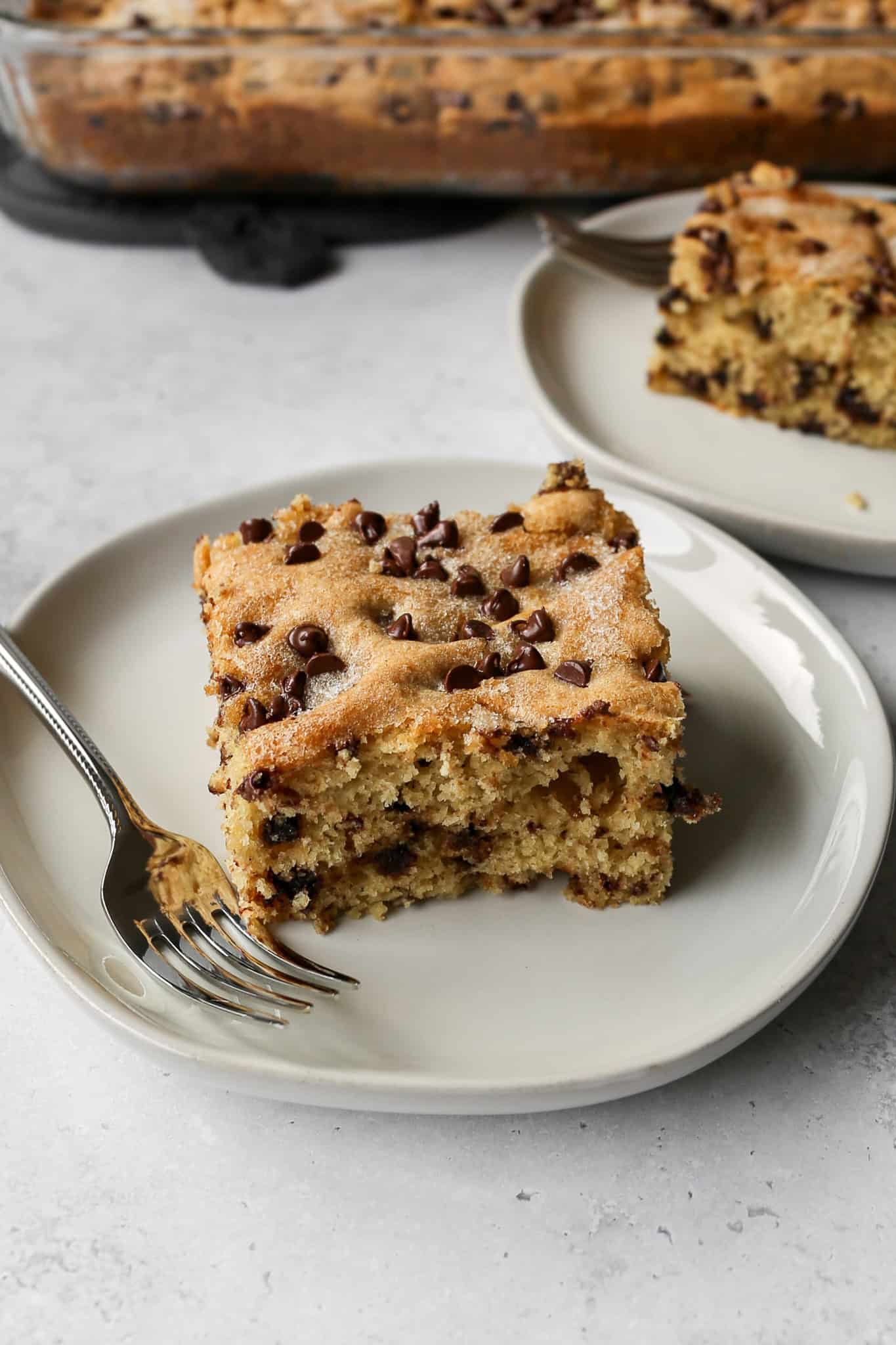 a square slice of gluten free chocolate chip snack cake on a white plate with a silver fork next to it