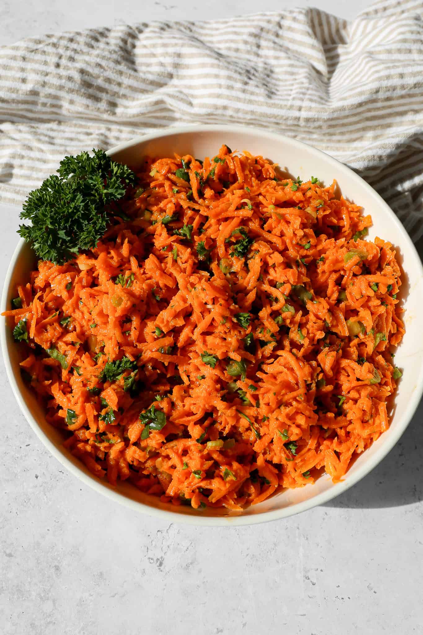 a cream serving bowl of honey dijon carrot salad with a parsley garnish next to a tan and white striped kitchen towel