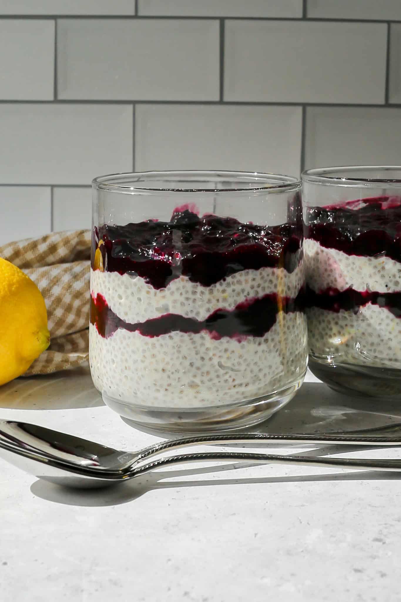 a glass of lemon blueberry chia pudding next to silver spoons, a lemon and tan gingham kitchen towel