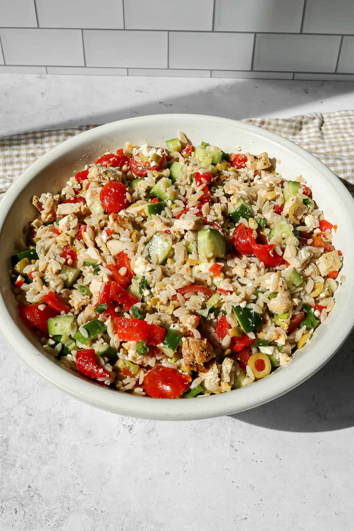 mediterranean chicken orzo salad in a white bowl next to a tan and white gingham kitchen towel