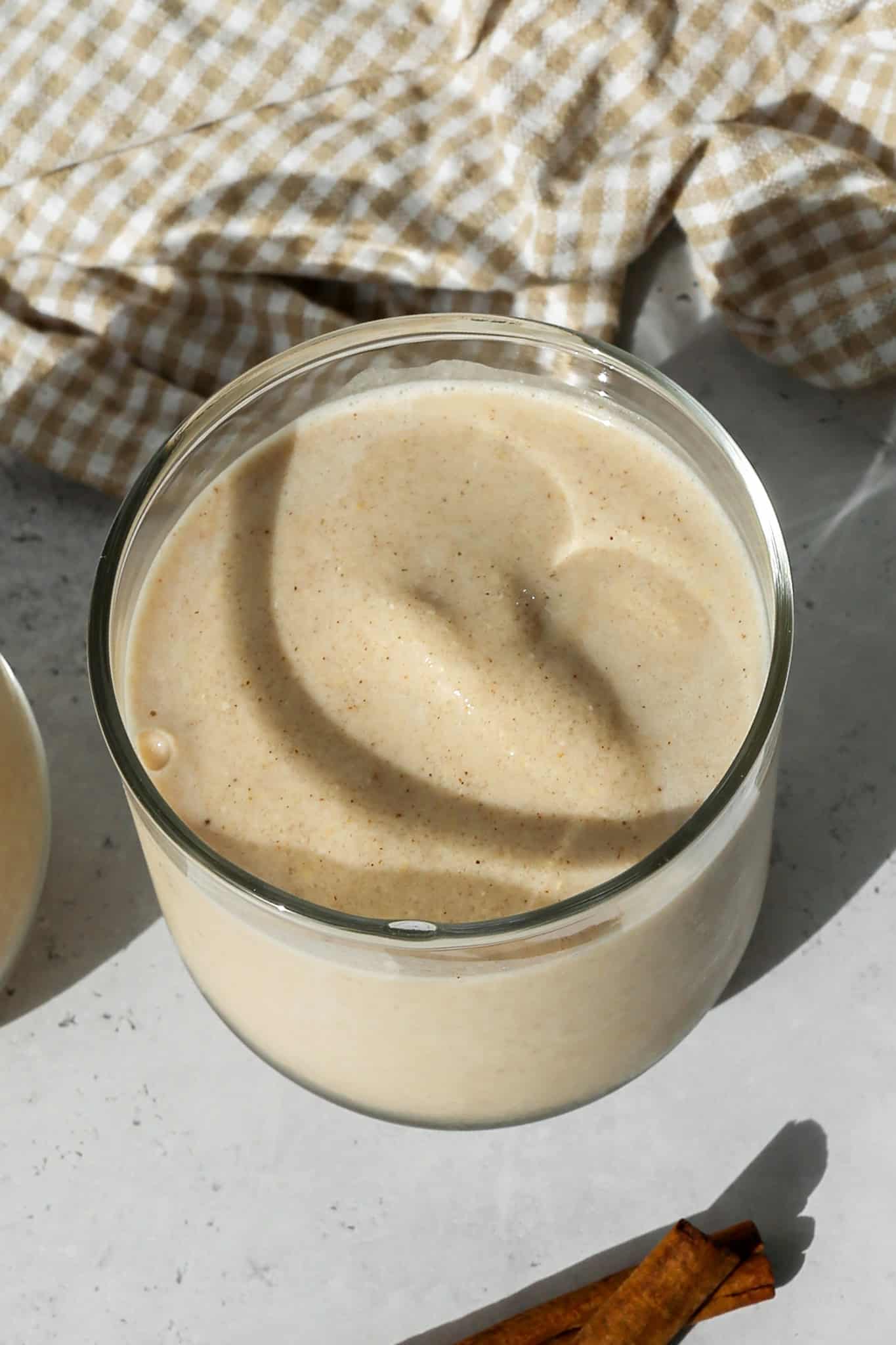 a clear glass of vanilla chai protein smoothie next to a cinnamon stick and a tan and white gingham kitchen towel