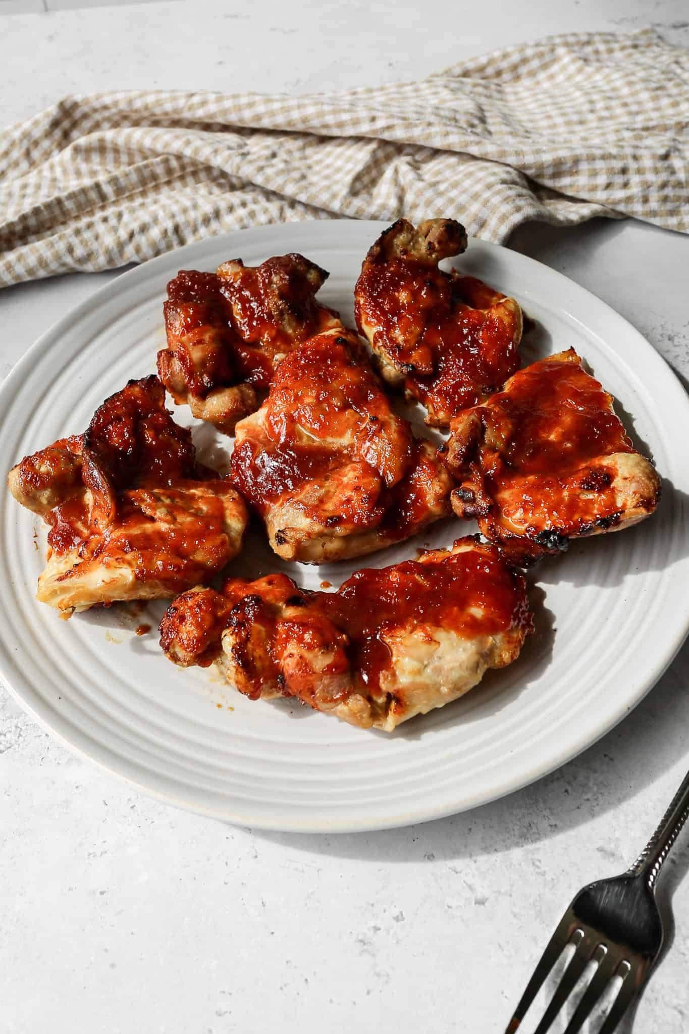 air fryer barbecue chicken thighs on a white plate next to a tan and white gingham kitchen towel