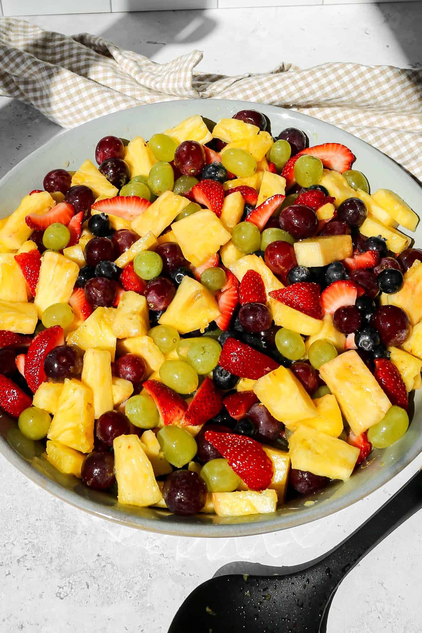 a large serving bowl of fruit salad with honey lime dressing nexts to a tan and white gingham kitchen towel