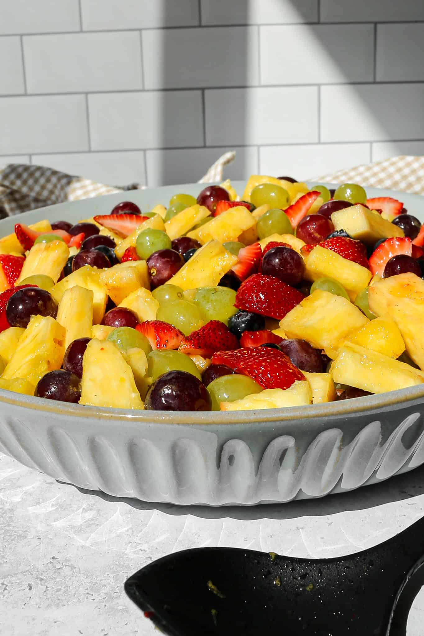 a large serving bowl of fruit salad with honey lime dressing nexts to a tan and white gingham kitchen towel