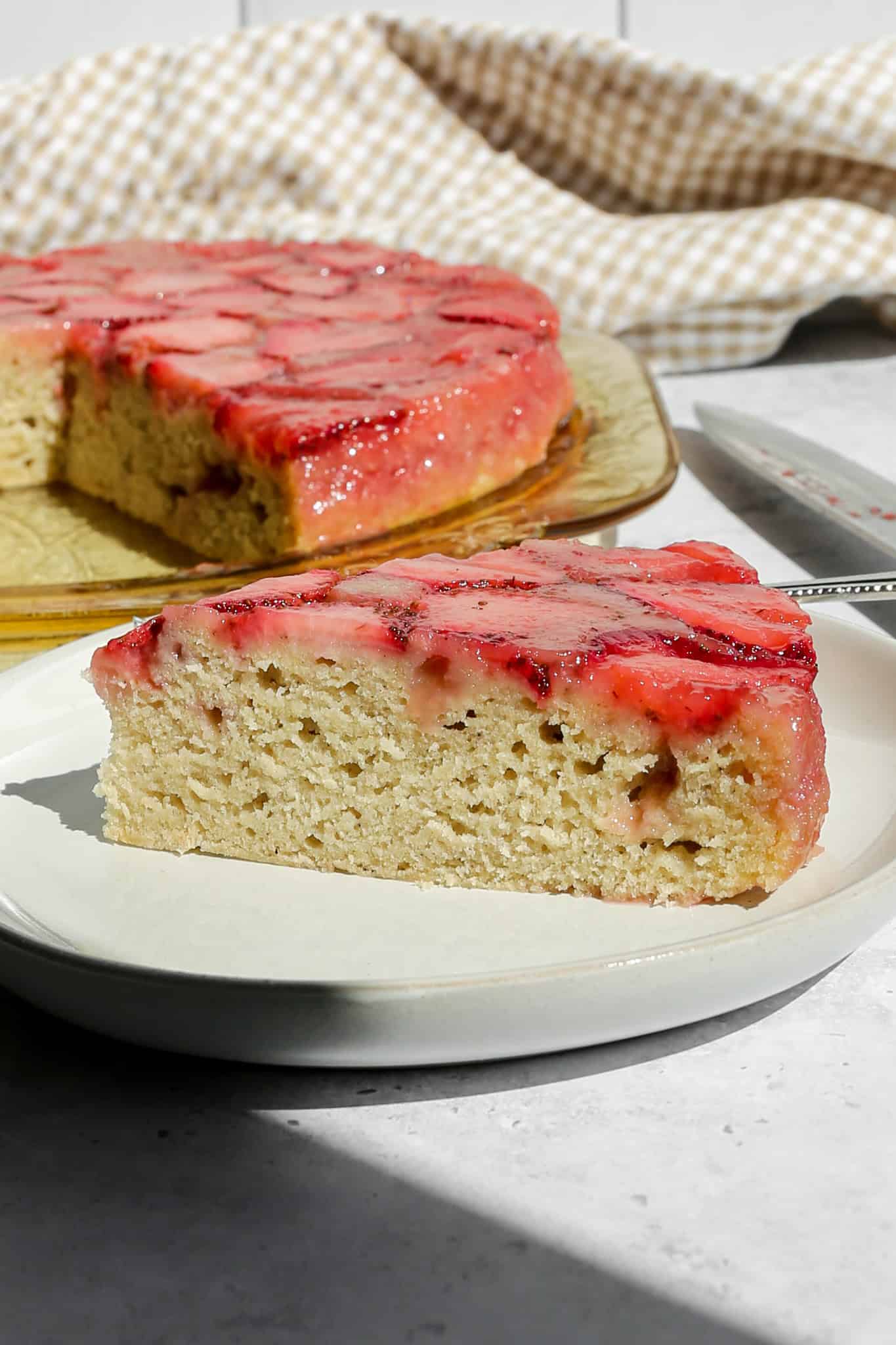 a slice of gluten free strawberry upside down cake on a beige plate next to the cut cake on a platter