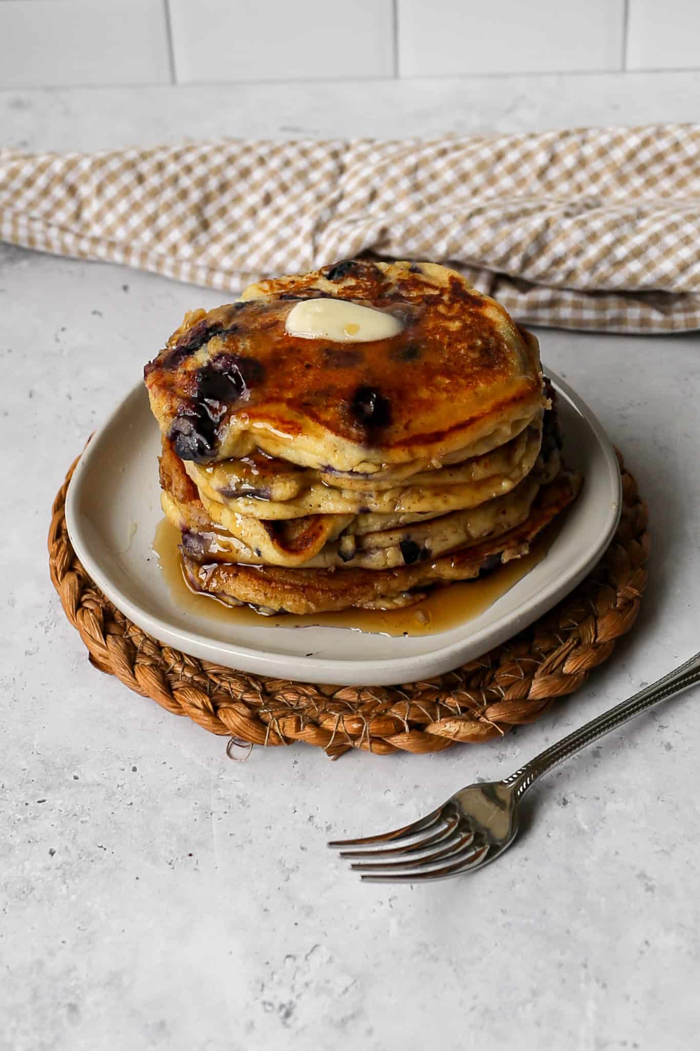 a white plate of five gluten free high protein lemon blueberry pancakes on a woven trivet next to a tan gingham kitchen towel