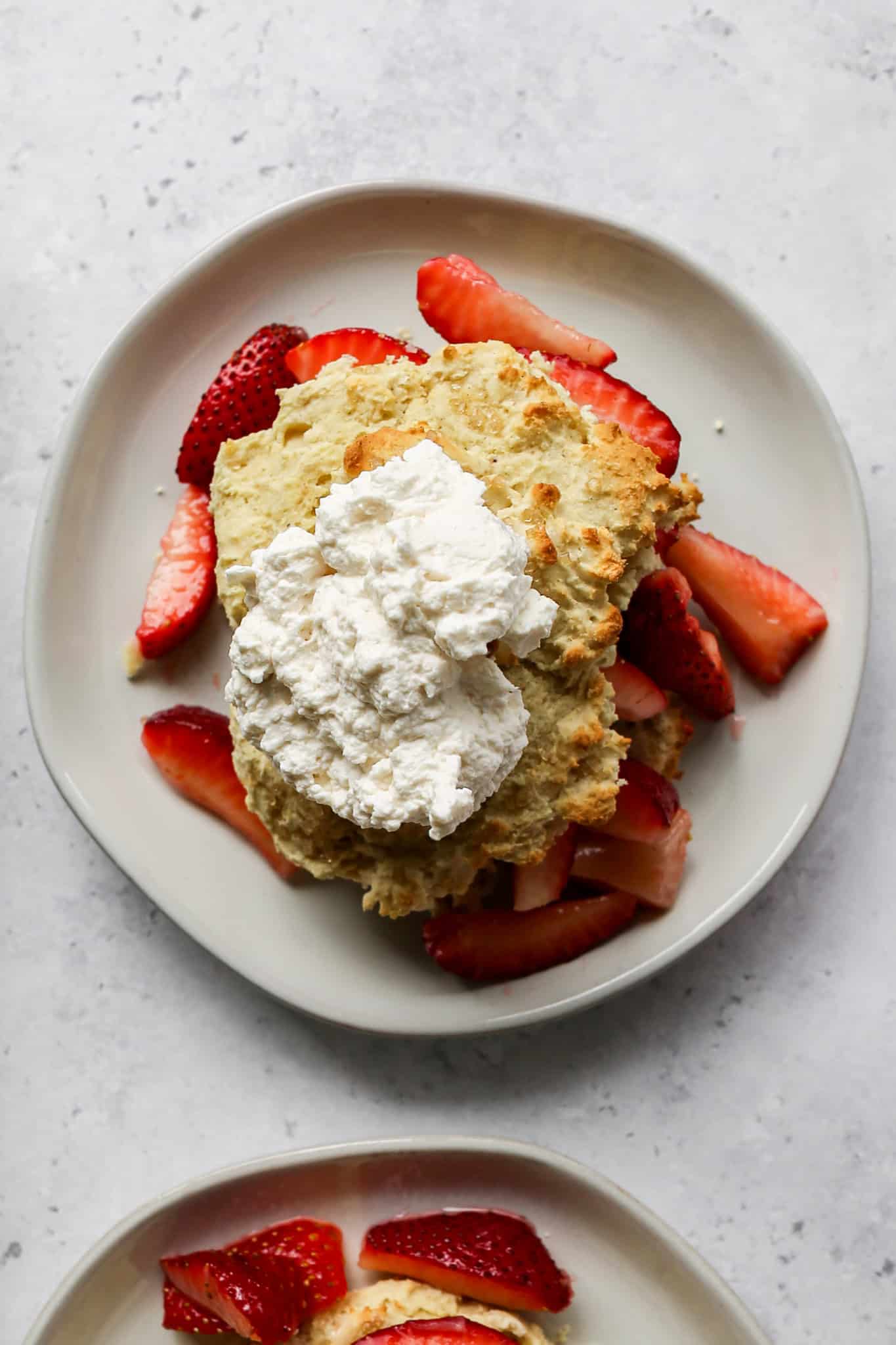 an off white plate with a gluten free shortcake biscuit cut in half with sliced strawberries on the inside and homemade whipped cream on top