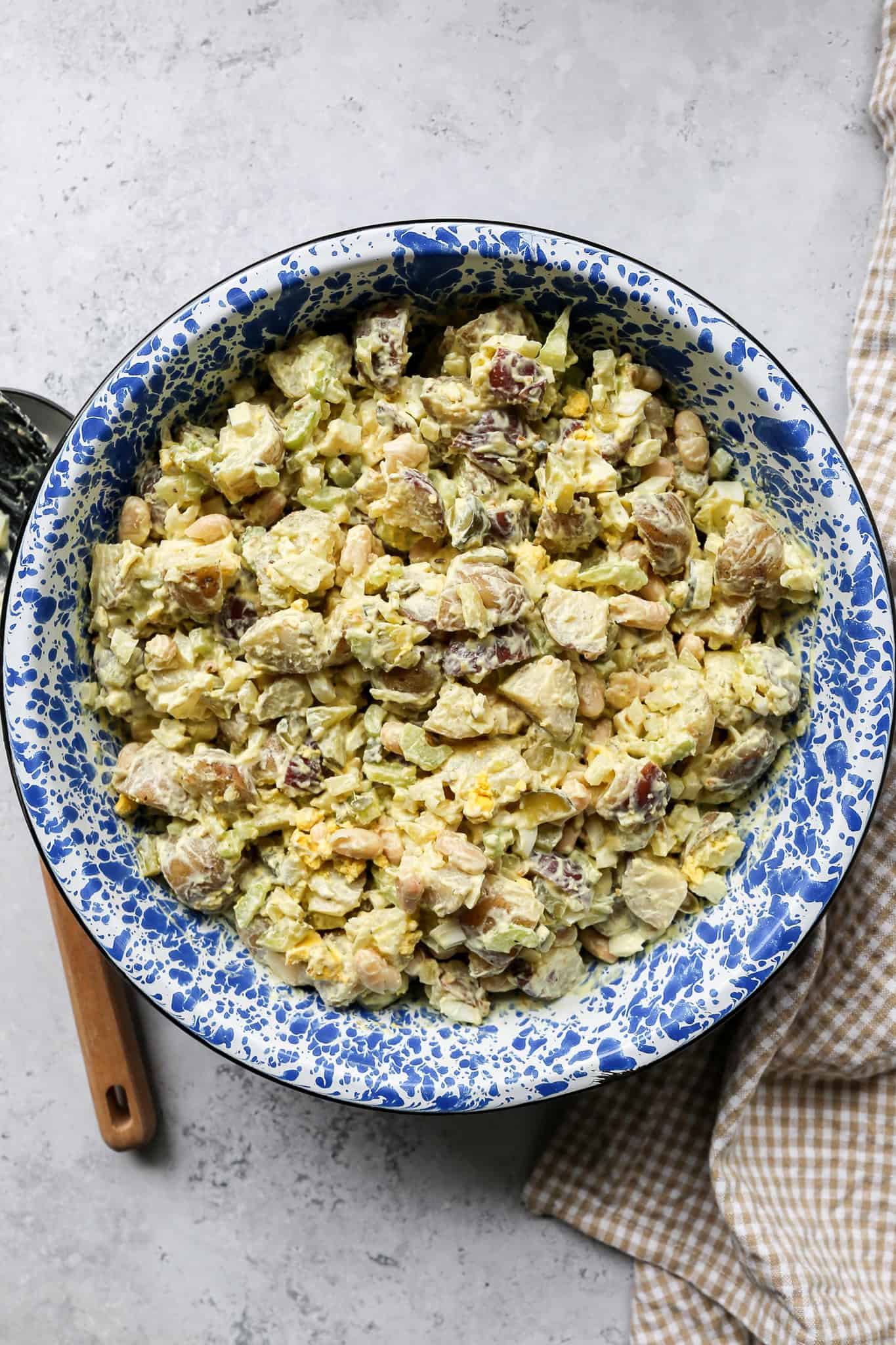 high protein potato salad in a blue enamelware bowl next to a tan gingham kitchen towel and a large serving spoon