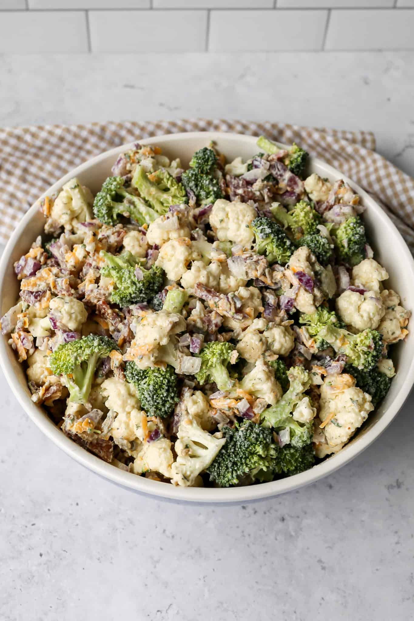 a bowl of high protein ranch broccoli cauliflower salad next to a tan and white gingham kitchen towel