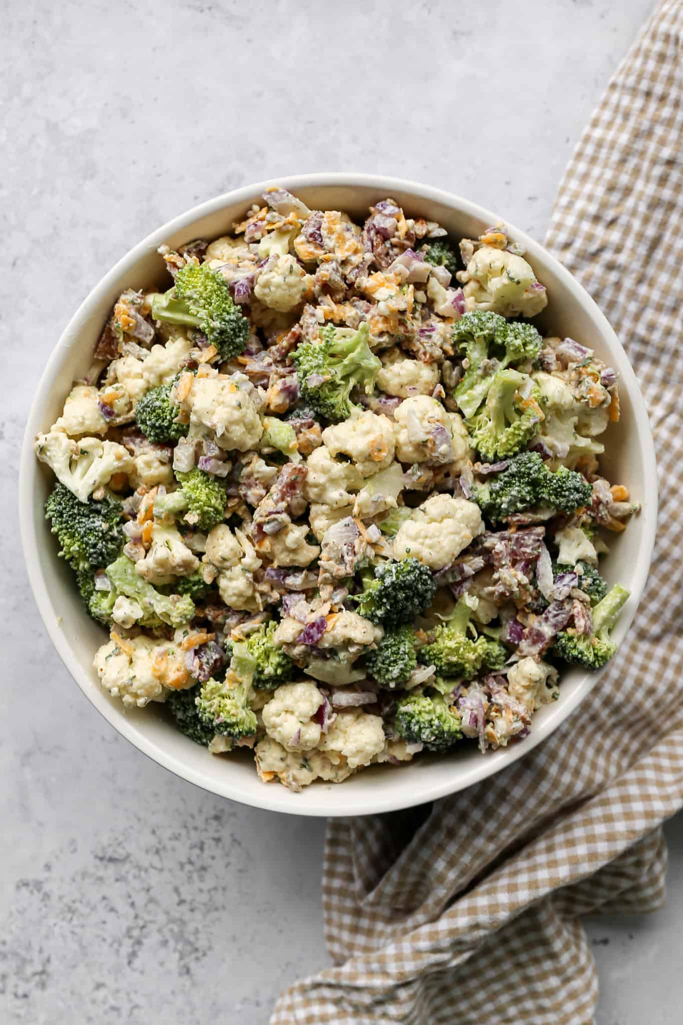 a bowl of high protein ranch broccoli cauliflower salad next to a tan and white gingham kitchen towel