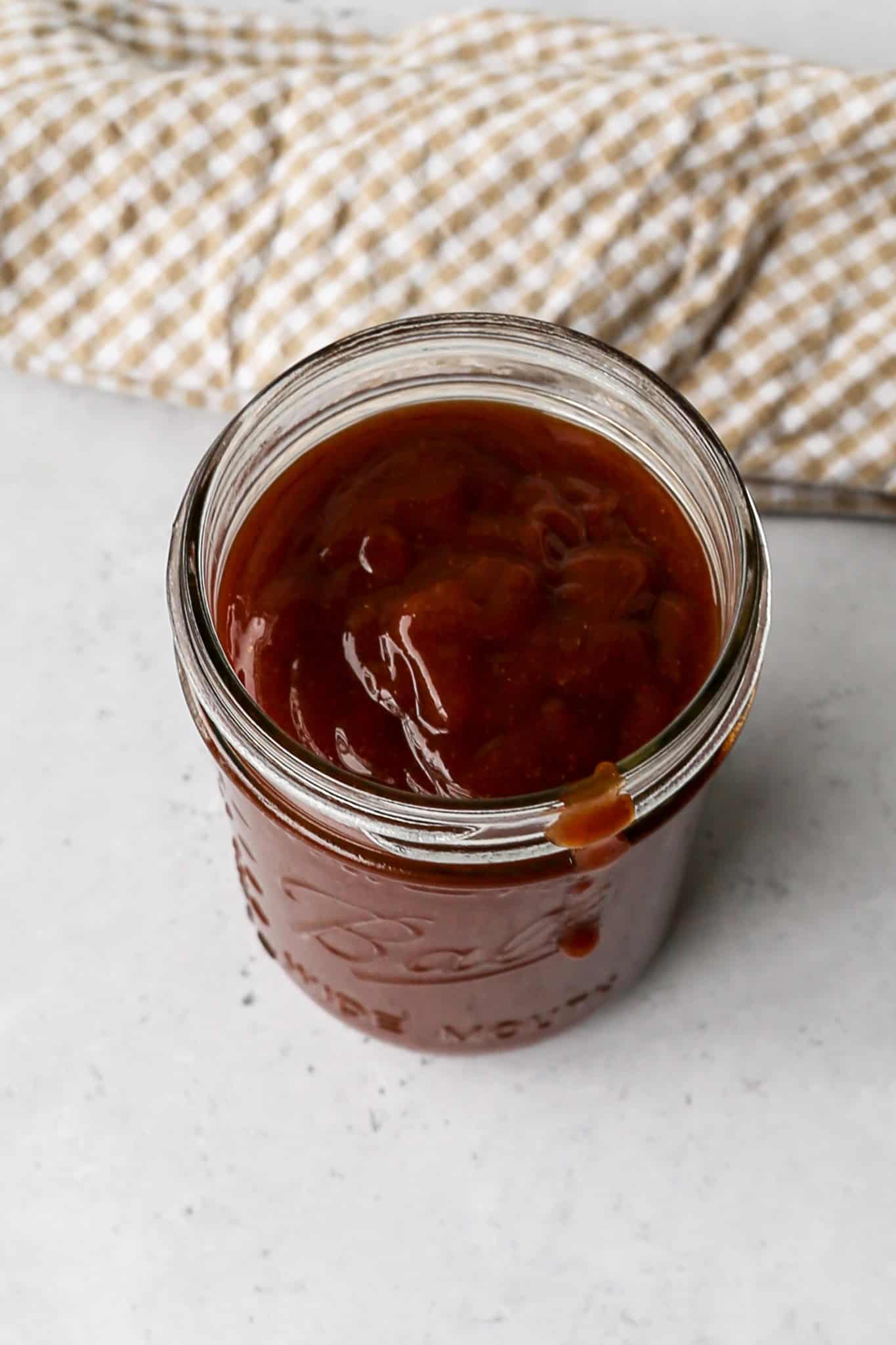 a wide mouth Ball canning jar of homemade BBQ sauce next to a tan and white gingham kitchen towel