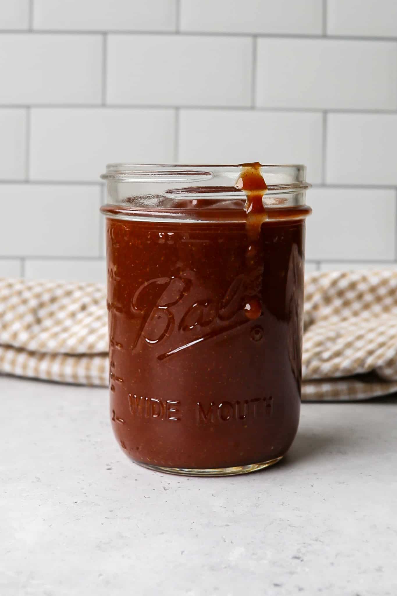 a wide mouth Ball canning jar of homemade BBQ sauce next to a tan and white gingham kitchen towel
