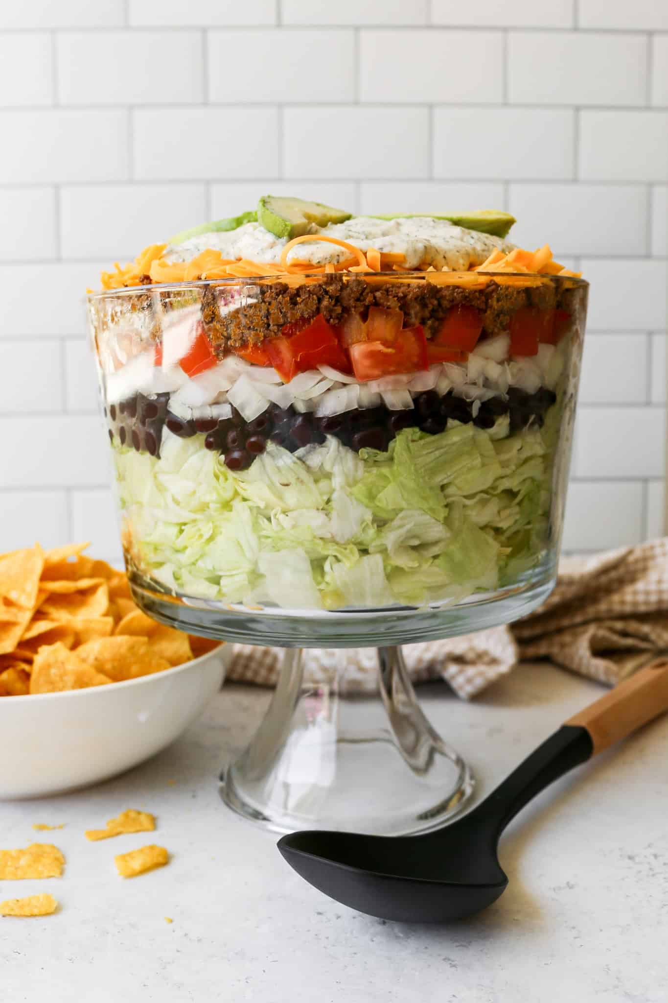 a clear trifle dish of layer taco salad next to a cream bowl of gluten free tortilla chips and a tan and white gingham kitchen towel