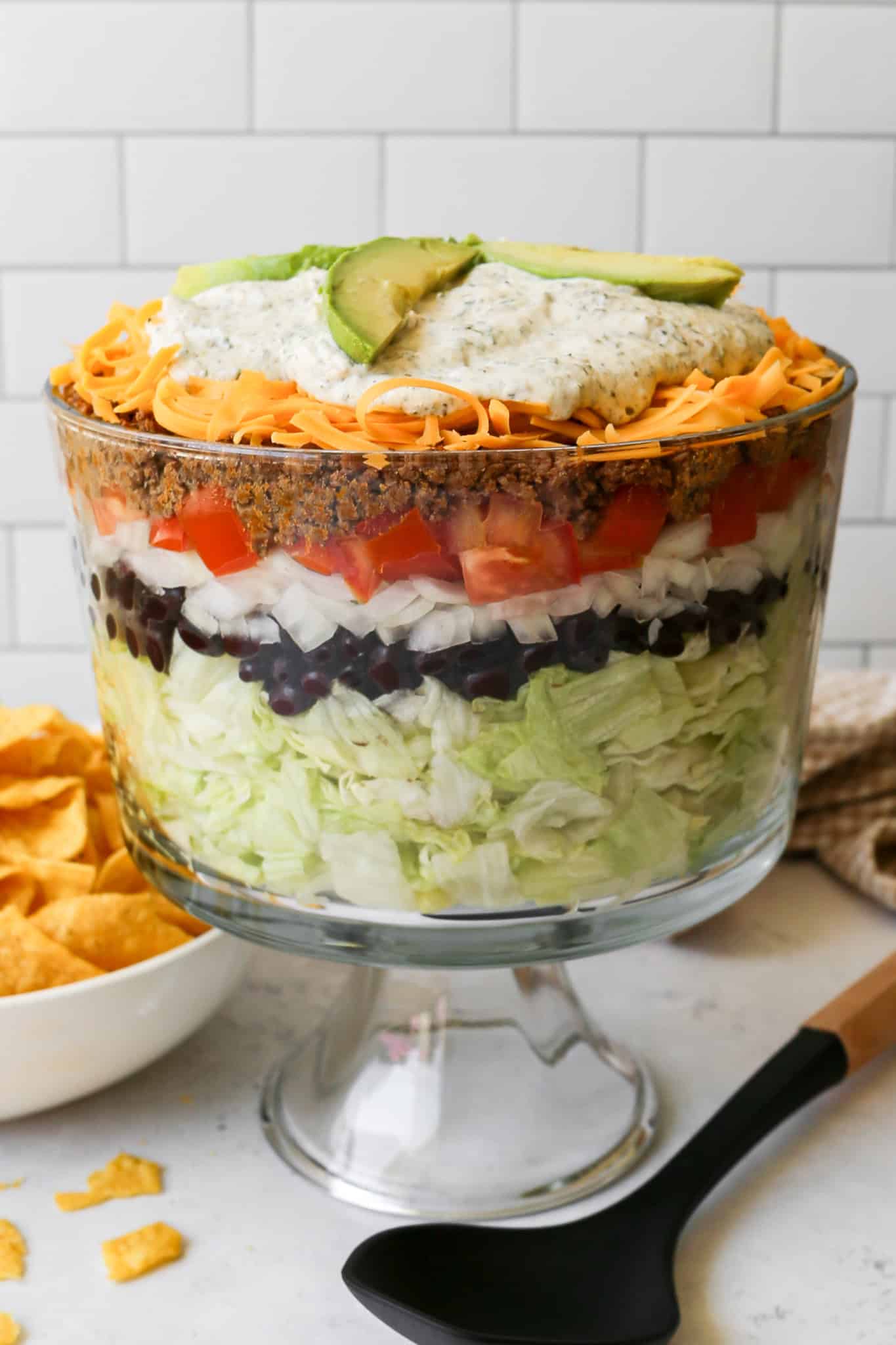 a clear trifle dish of layer taco salad next to a cream bowl of gluten free tortilla chips and a tan and white gingham kitchen towel