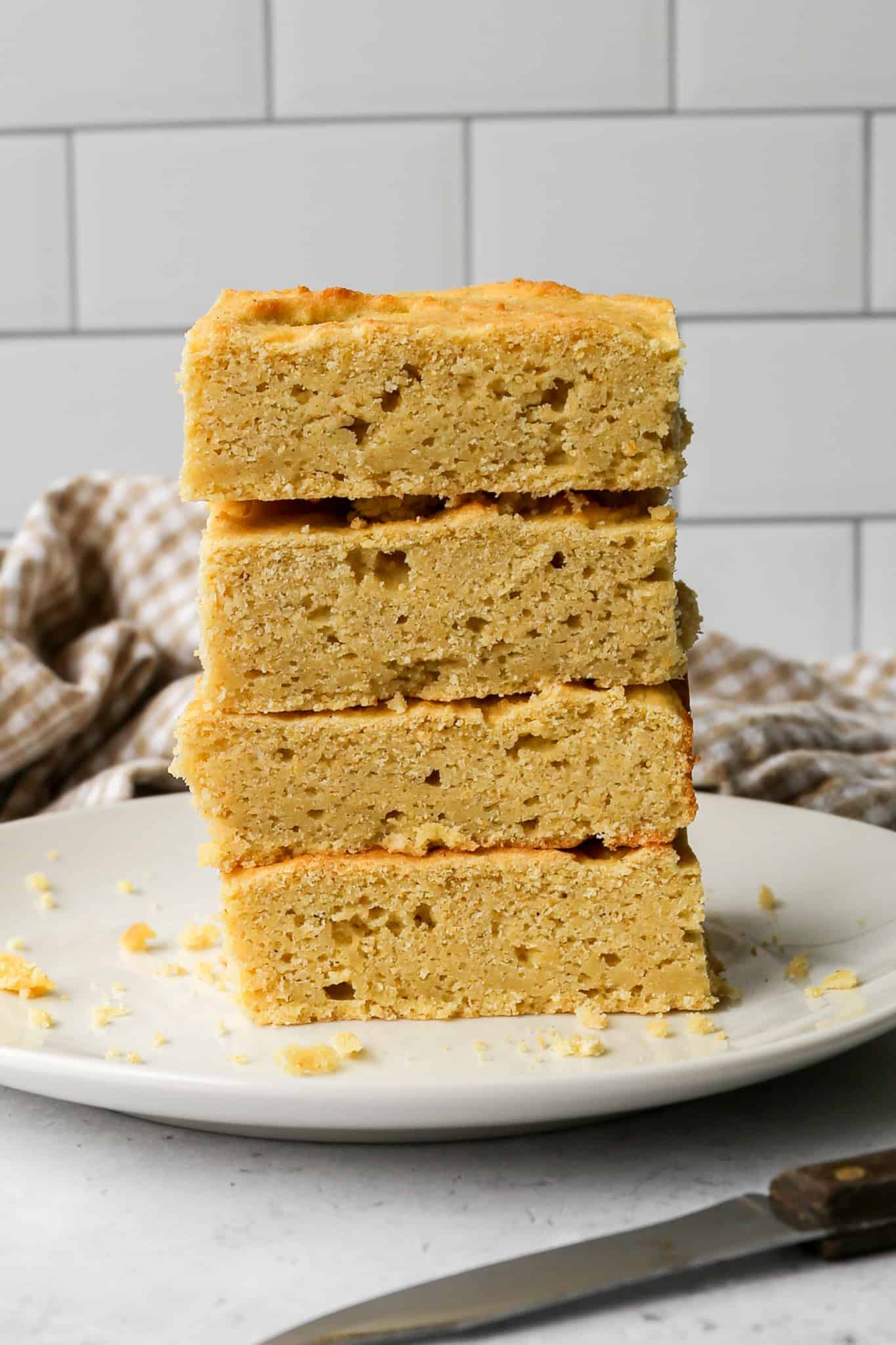 a stack of four pieces of gluten free buttermilk cornbread on a cream plate next to a tan gingham kitchen towel