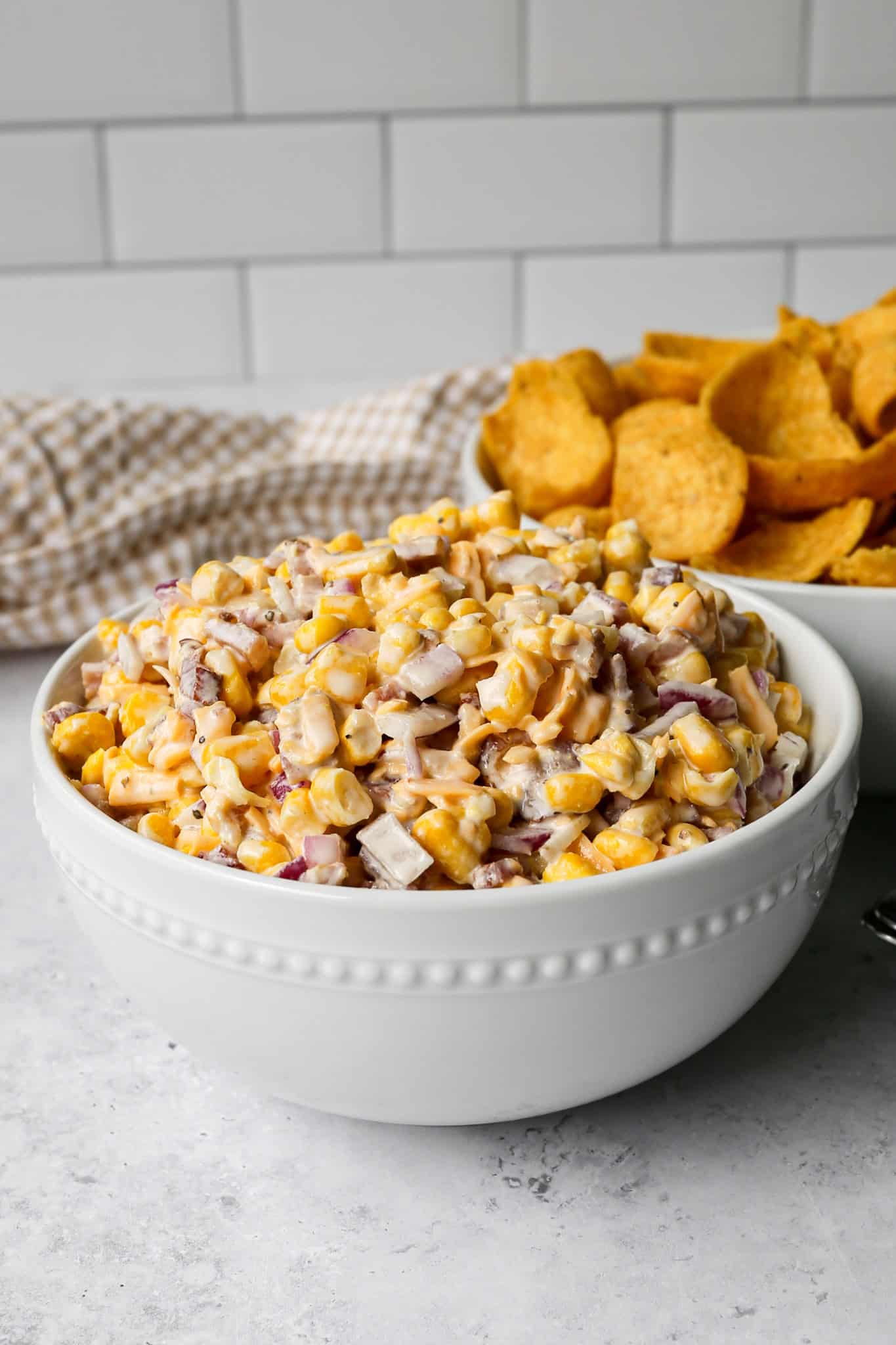 a white bowl of gluten free corn salad with bacon next to a white bowl of corn chips and a tan gingham kitchen towel