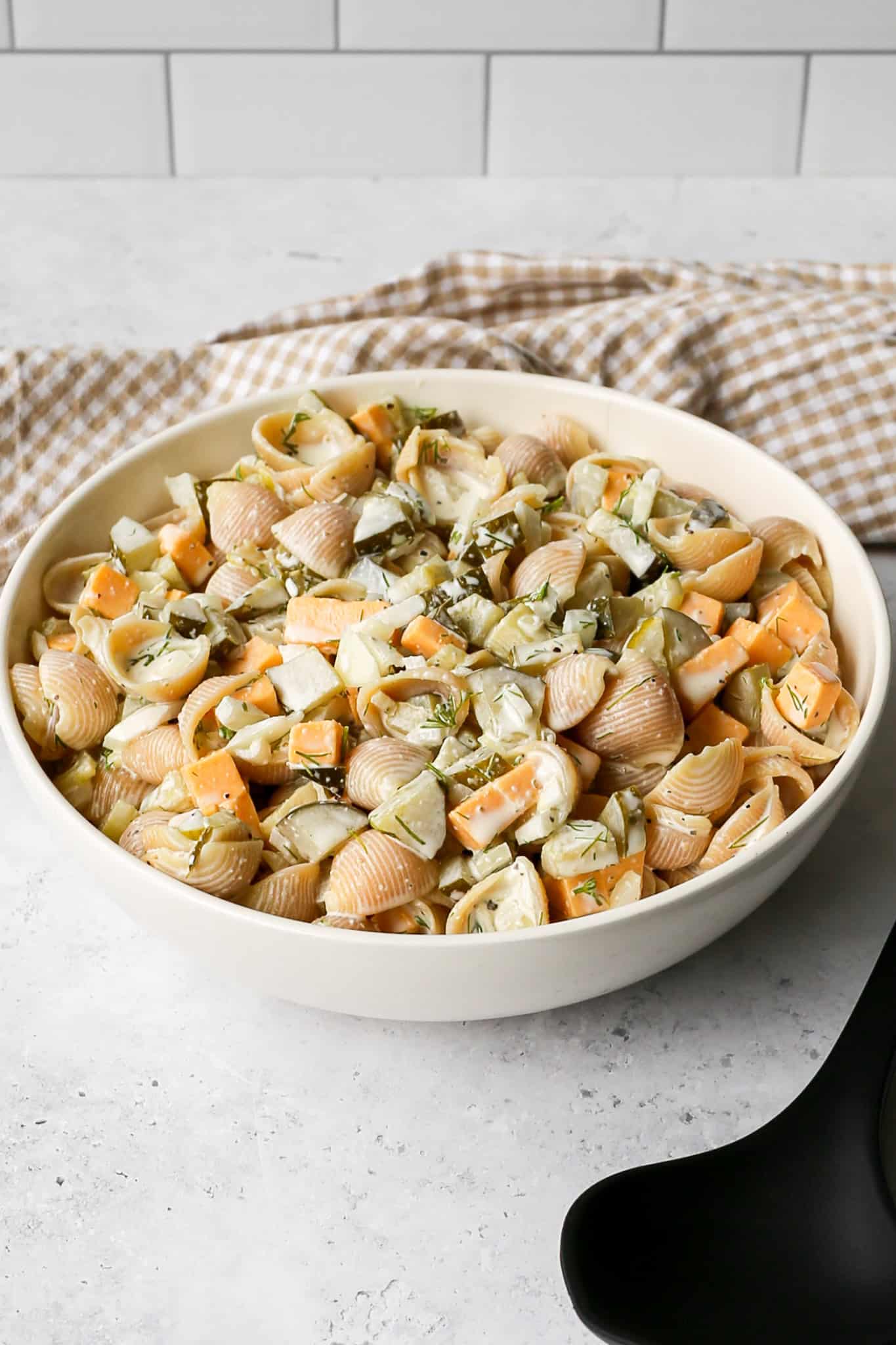 a cream serving bowl of gluten free dill pickle pasta salad next to a black serving spoon and tan gingham kitchen towel