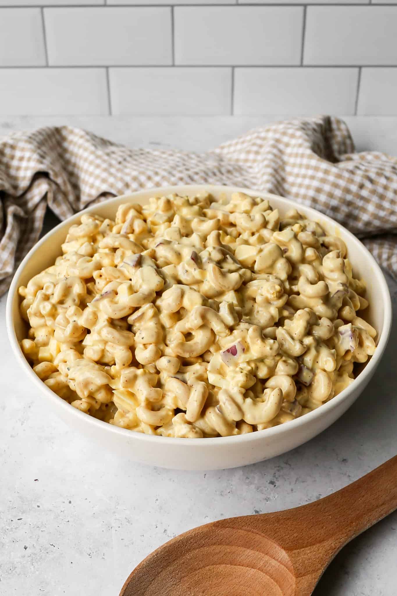 a bowl of gluten free high protein deviled egg pasta salad next to a wooden spoon and a tan gingham kitchen towel