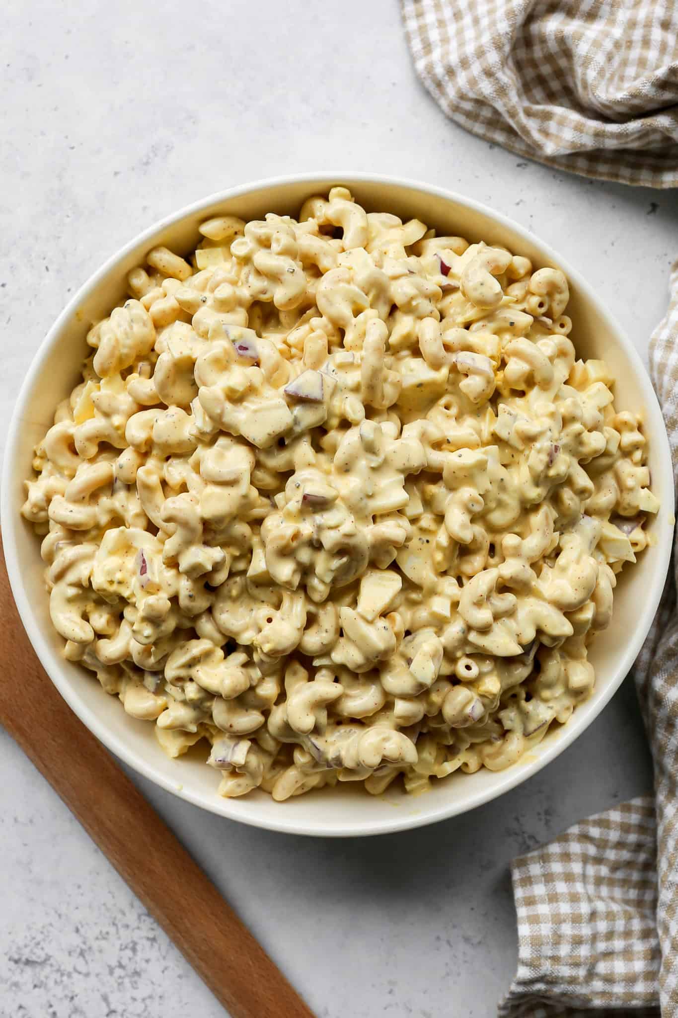 a bowl of gluten free high protein deviled egg pasta salad next to a wooden spoon and a tan gingham kitchen towel