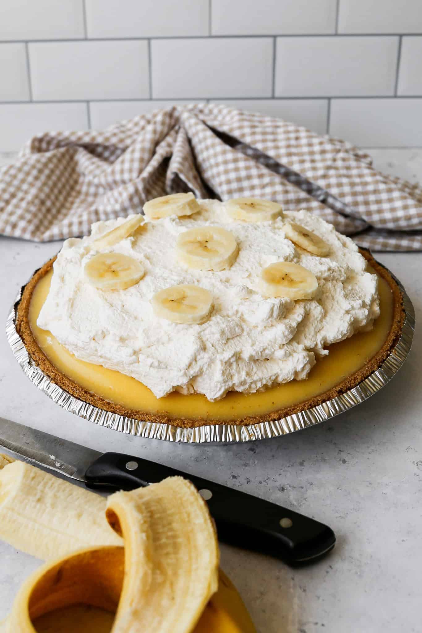 a gluten free banana cream pie in a silver pie pan next to a banana, a knife and a tan gingham kitchen towel