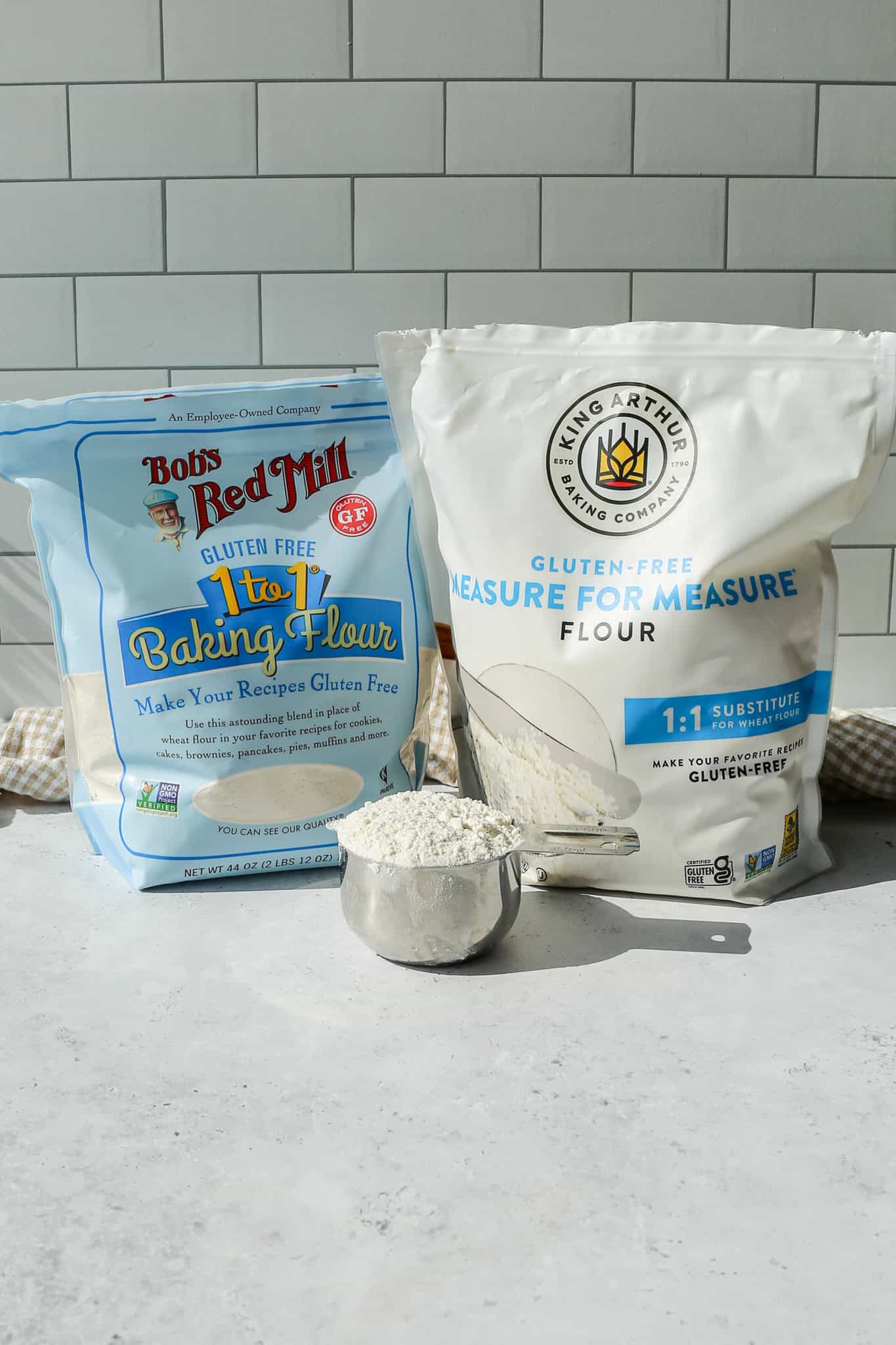 a bag of Bob's Red Mill Gluten Free 1 to 1 Baking Flour next to a bag of King Arthur Gluten Free Measure for Measure Flour with a silver measuring cup of flour in front of the bags